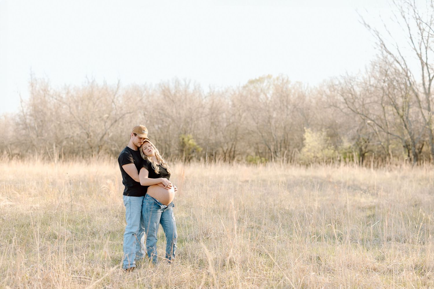A couple stands together in a serene autumn field with bare trees and golden grass in the background.