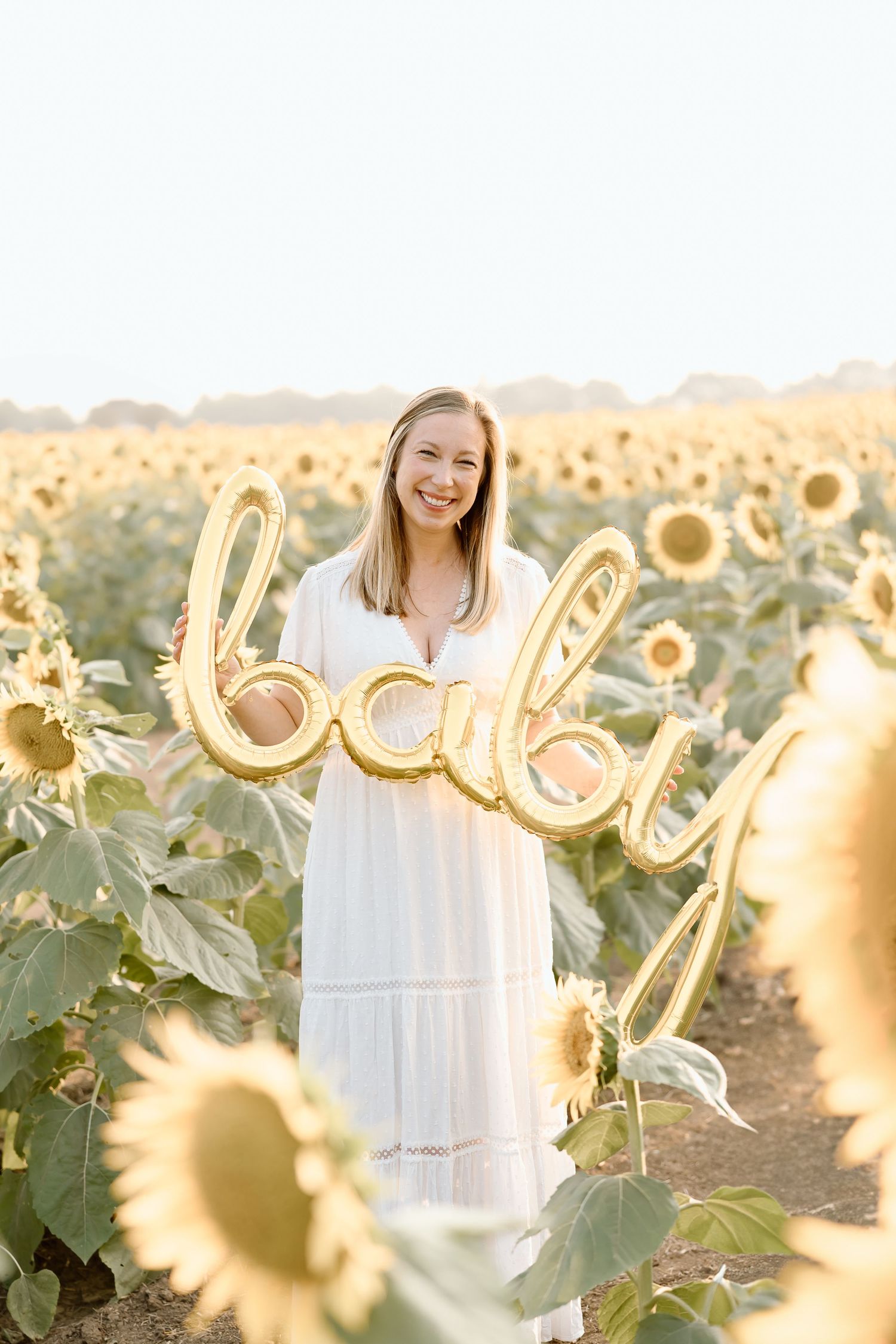 A person in a white dress stands joyfully among blooming sunflowers at sunset in a golden field.