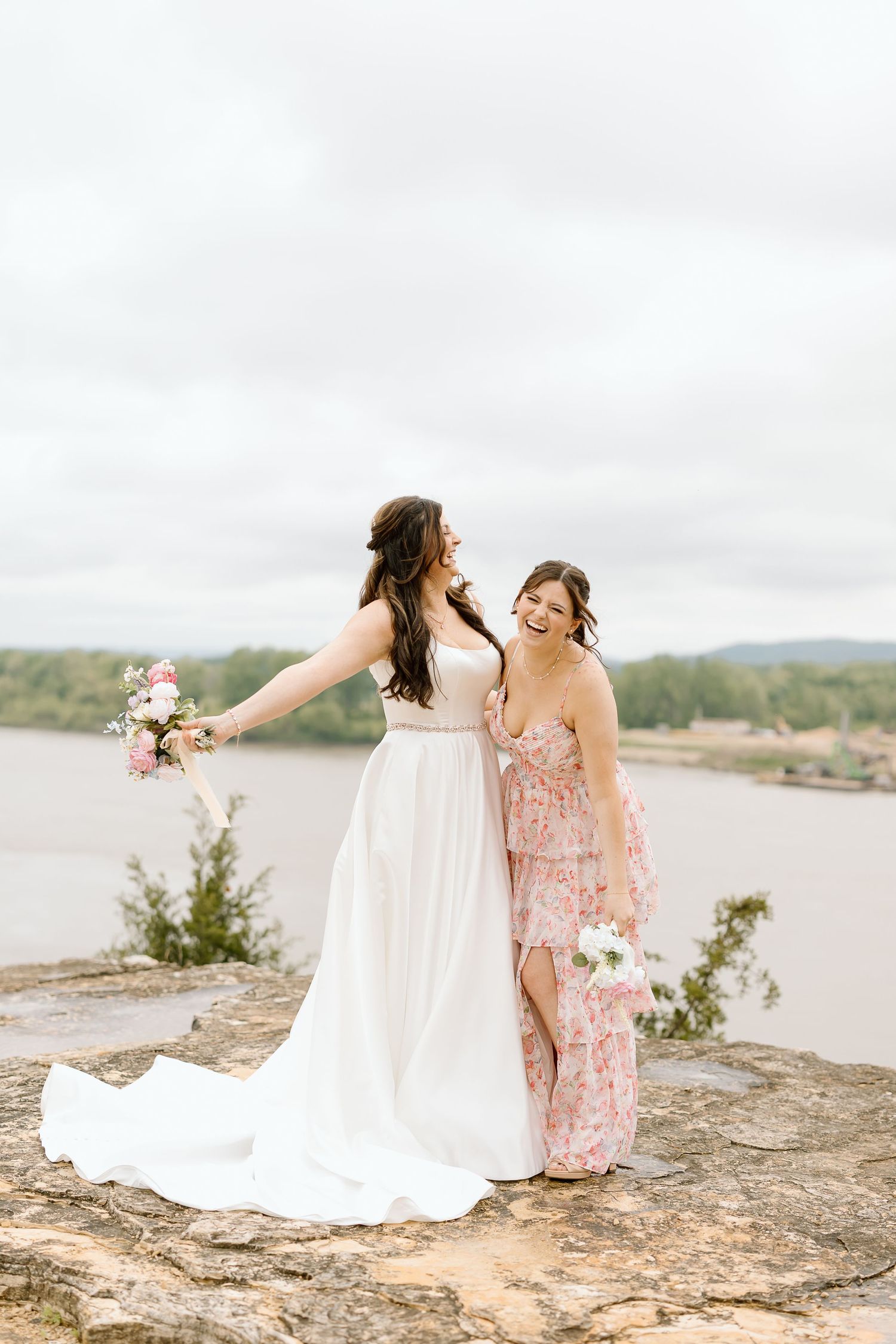 Two people in pink and white dresses sit together on a rocky overlook with a scenic landscape view in the background.