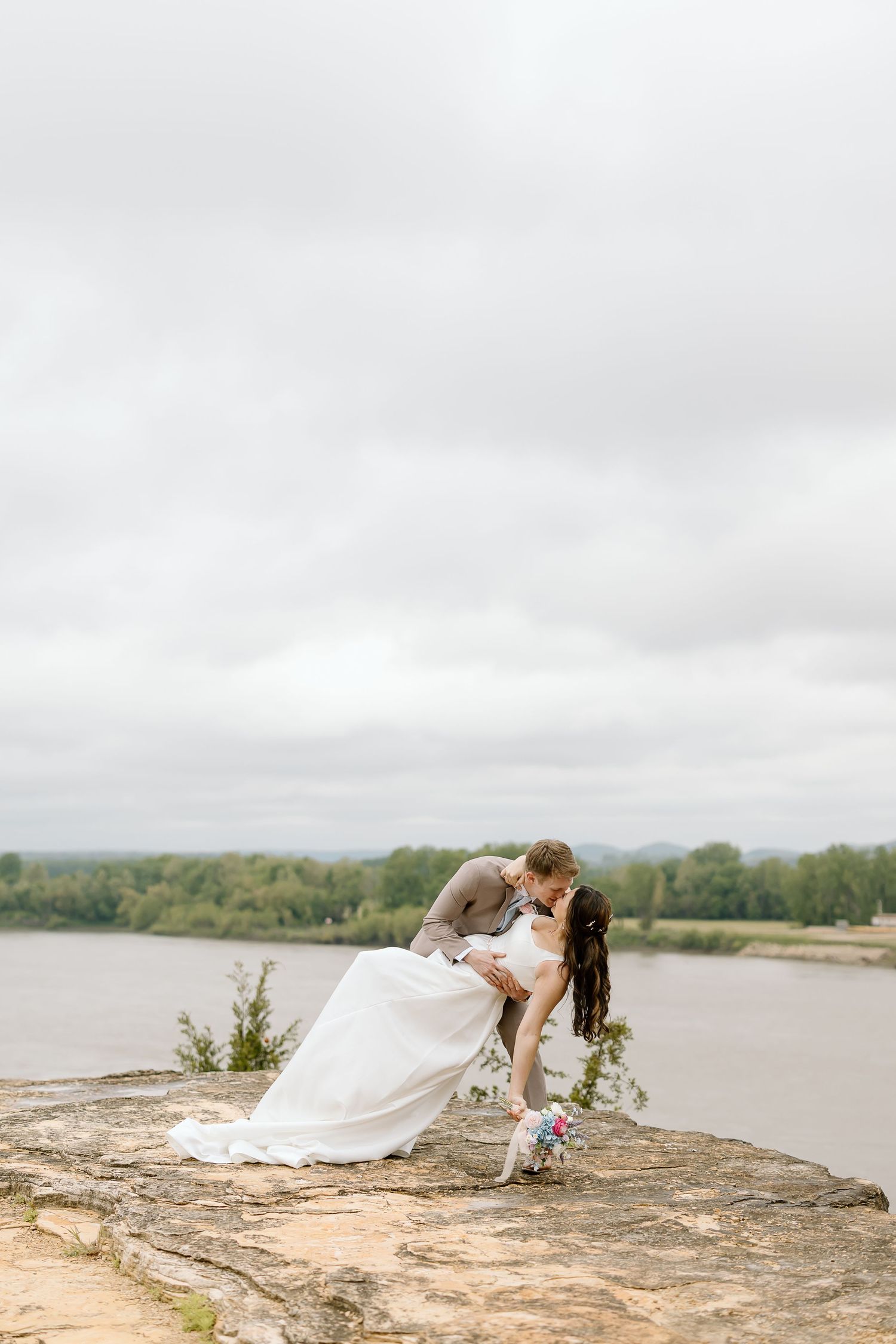 A romantic wedding couple shares intimate poses on a stone ledge overlooking a scenic lake view.