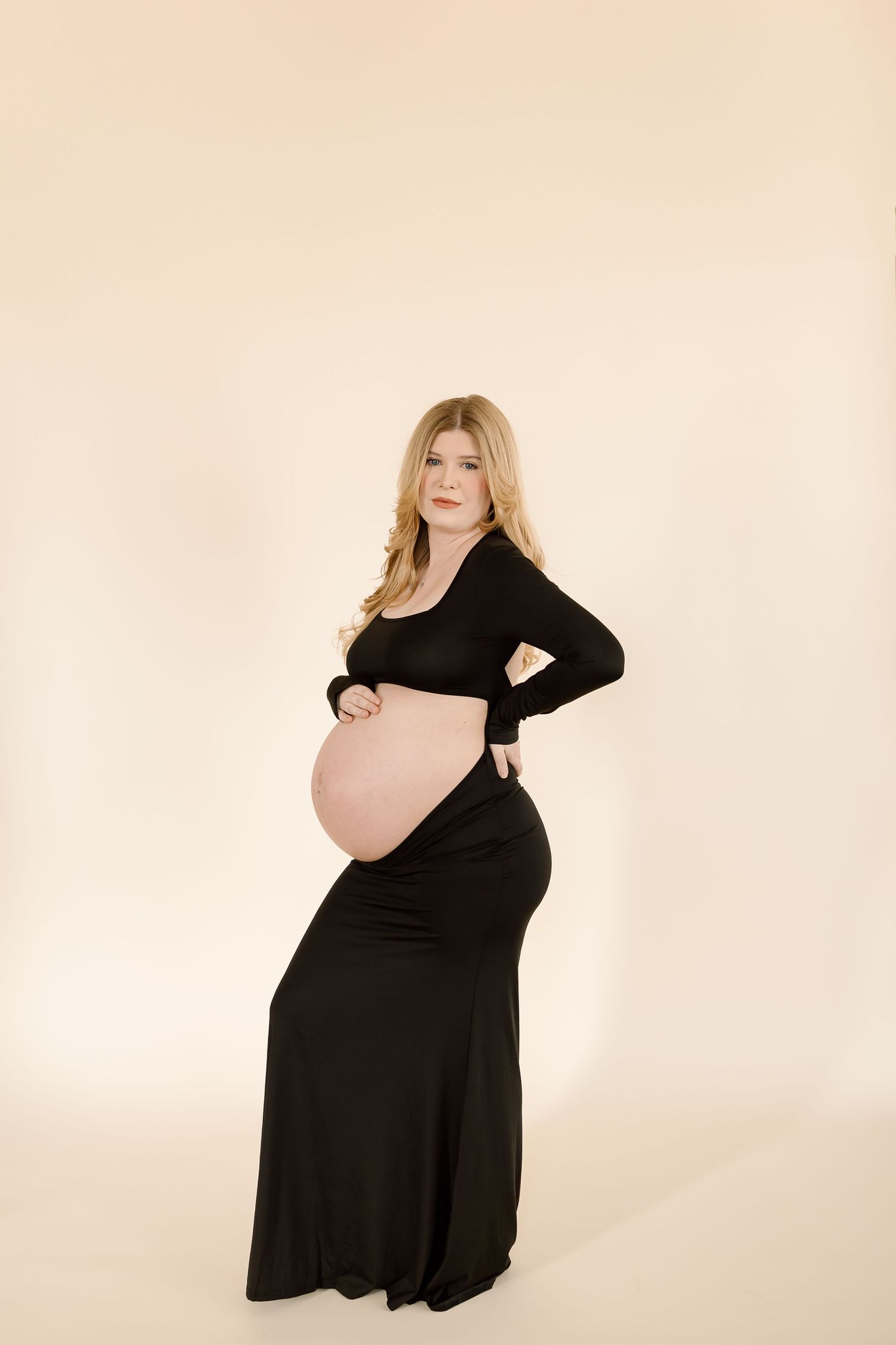 Expectant woman poses in a long black maternity dress against beige backdrop in elegant studio portrait.