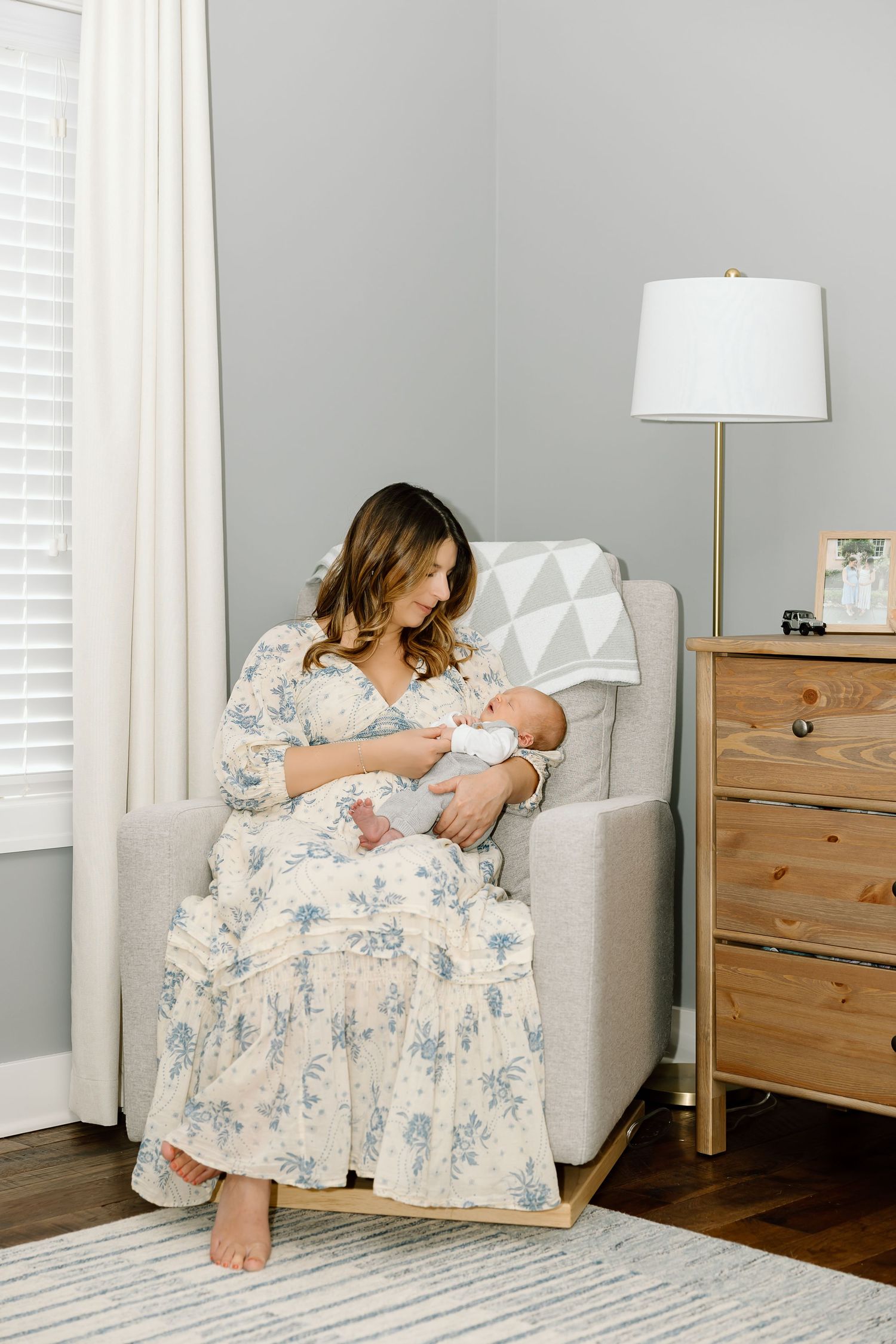 A mother in a floral dress sits in a gray armchair with a baby in a cozy bedroom setting with wooden furniture.