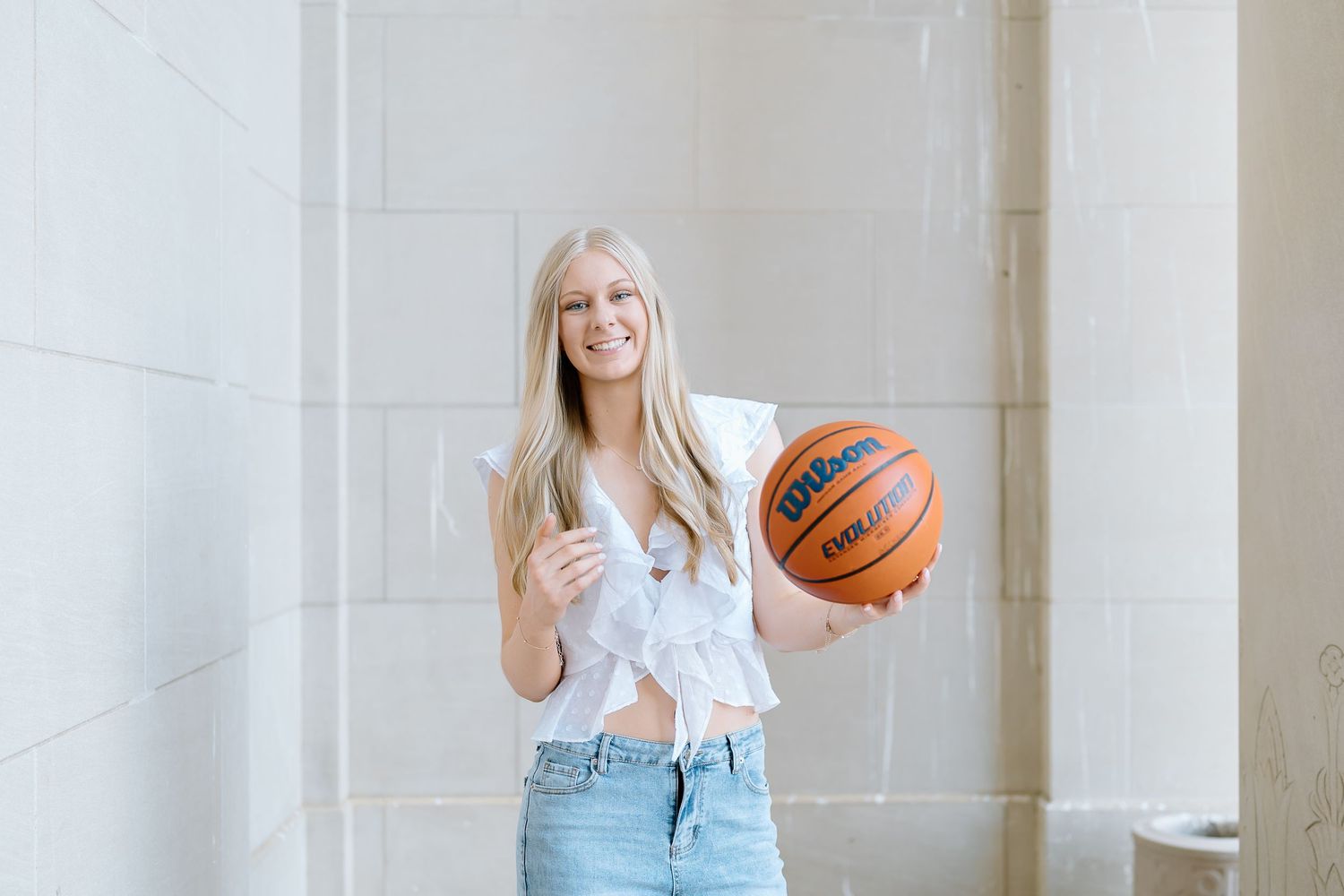 Person in light blue jeans and white top holding a basketball while standing against a white wall.