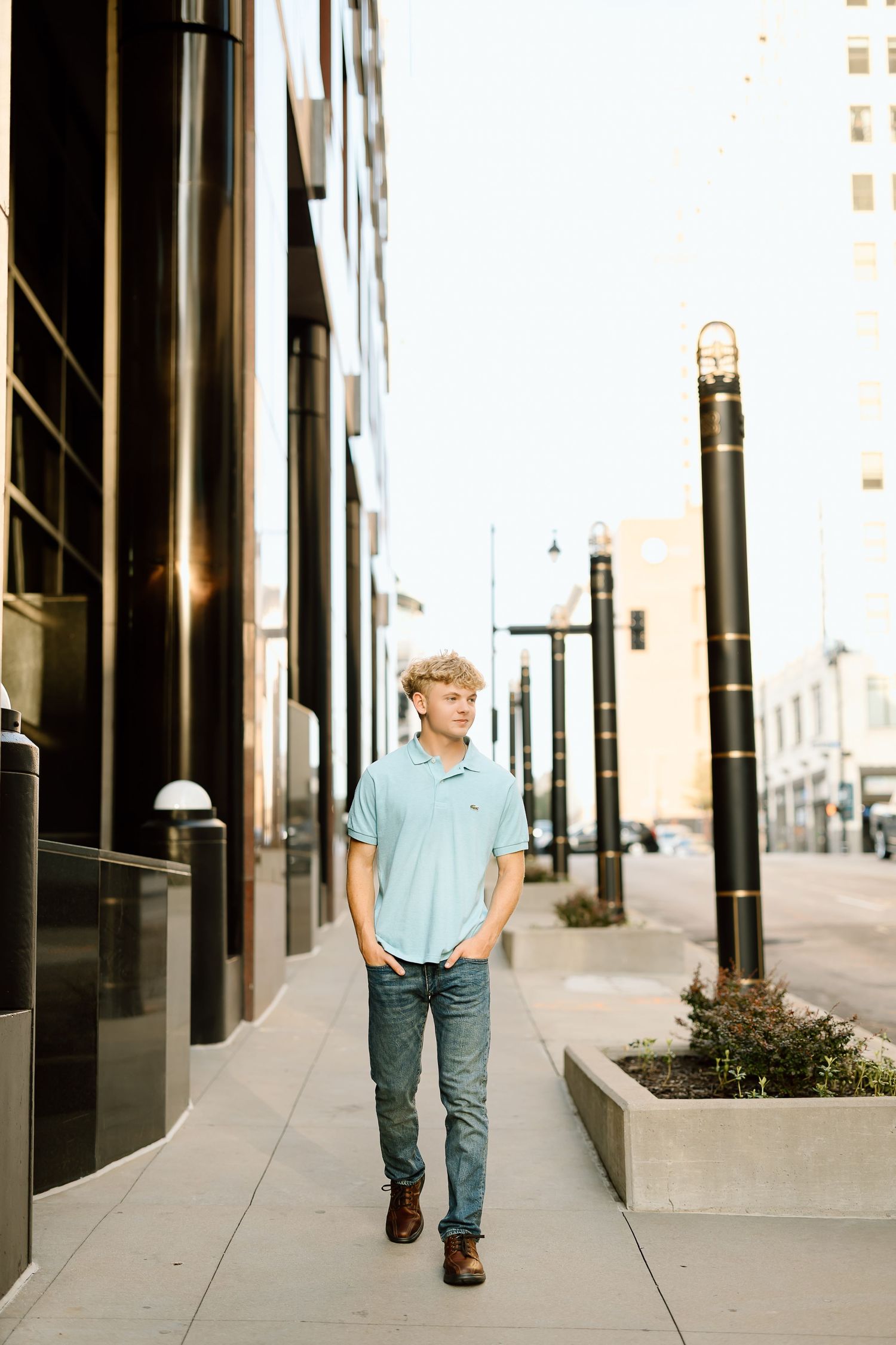 Person in light blue shirt and jeans walking down an urban sidewalk with black columns and modern architecture.