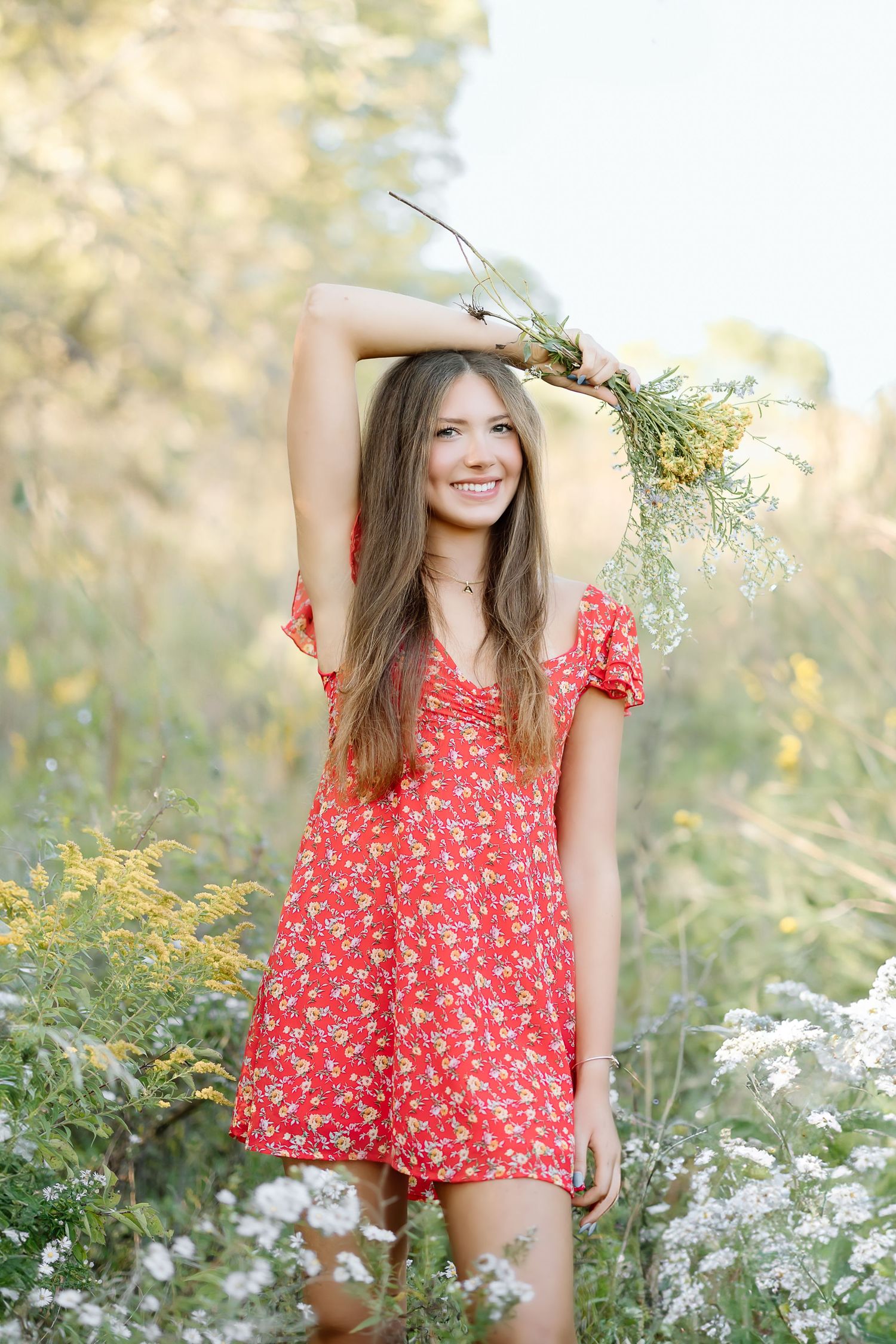 A person in a red floral romper poses playfully in a sunlit meadow with white wildflowers and natural greenery.