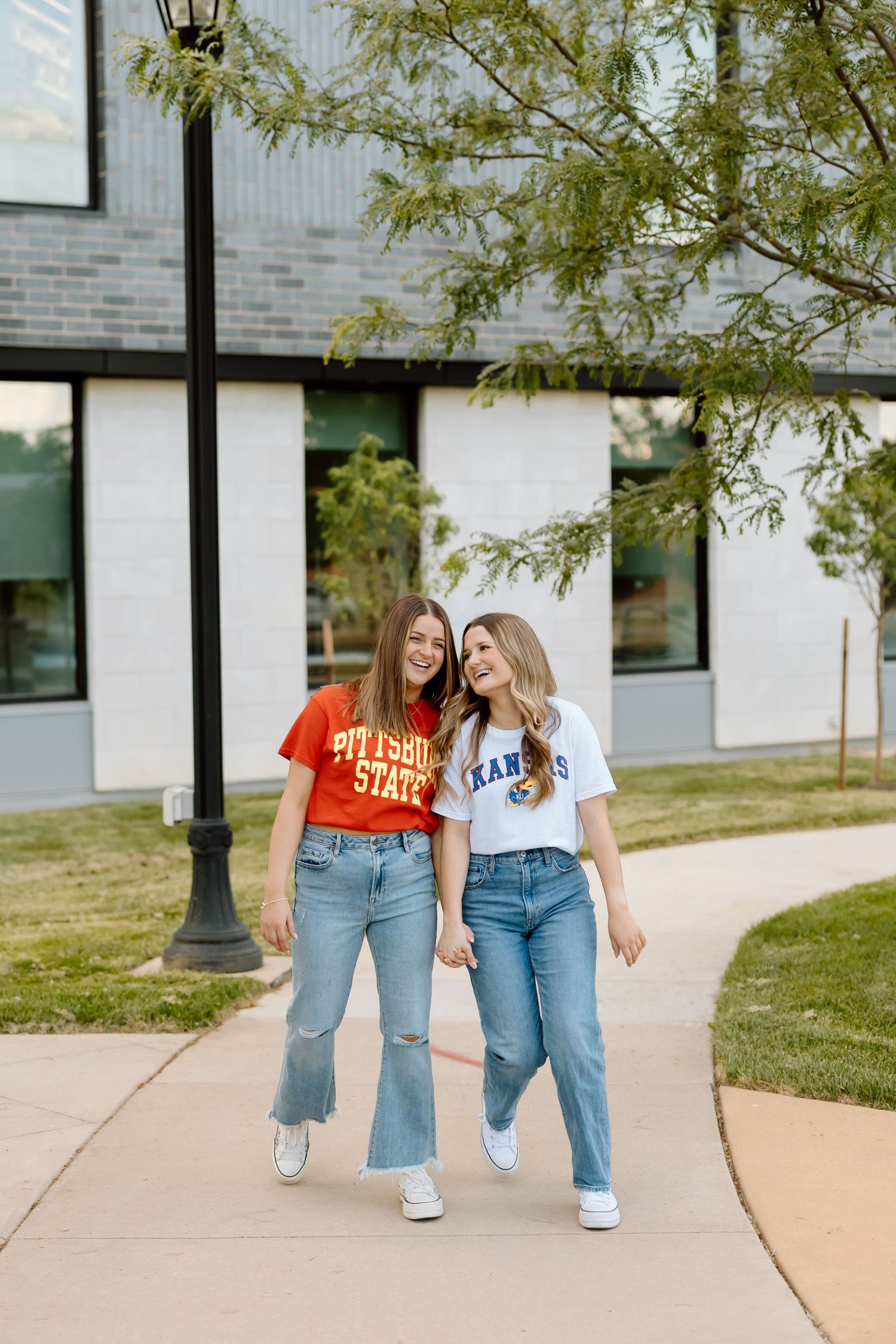 Two friends in matching jeans and sneakers walk together on a campus sidewalk on a sunny day.