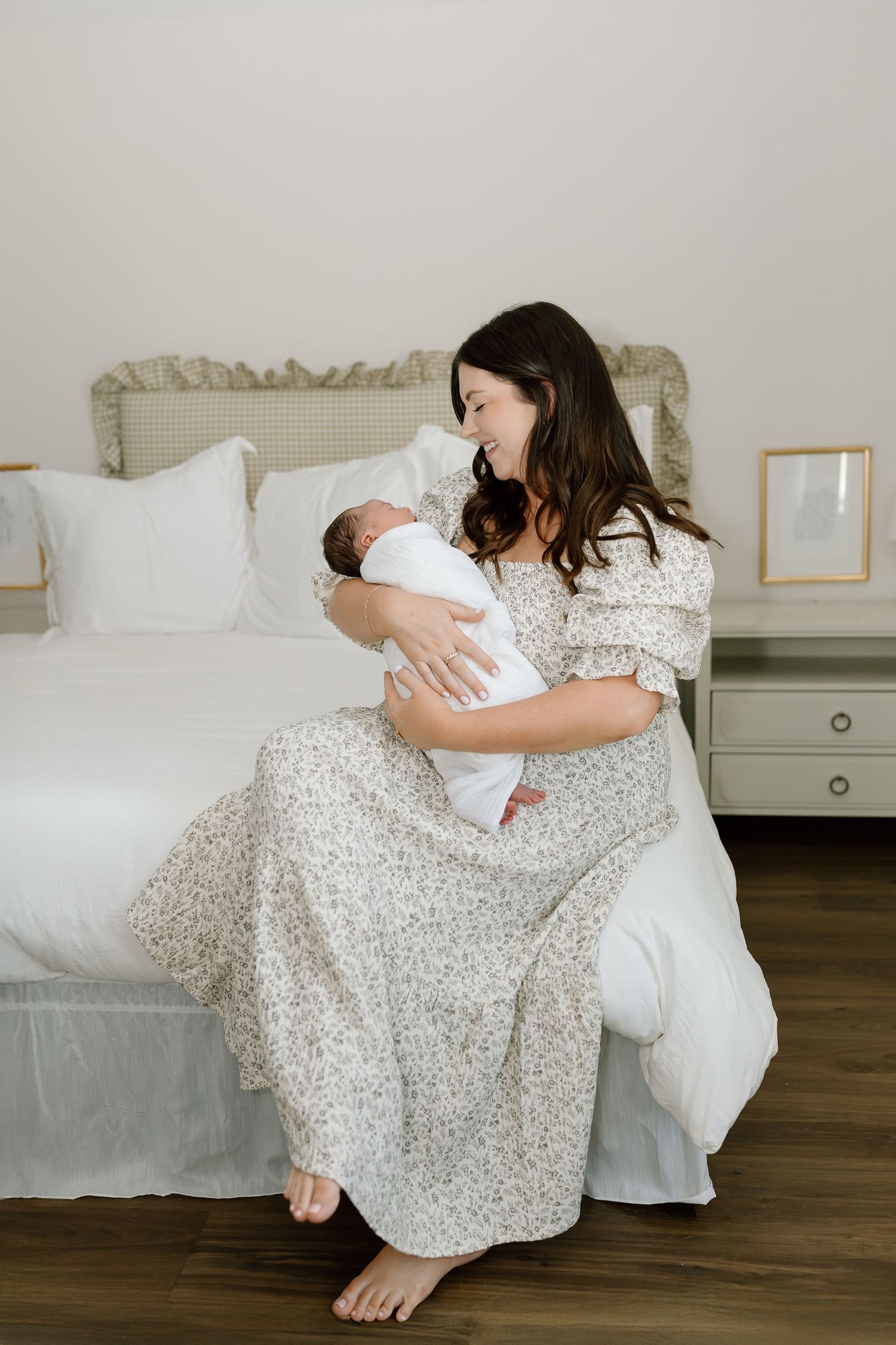 A mother in a floral dress sits on a hardwood floor cradling and bonding with her newborn baby in white.