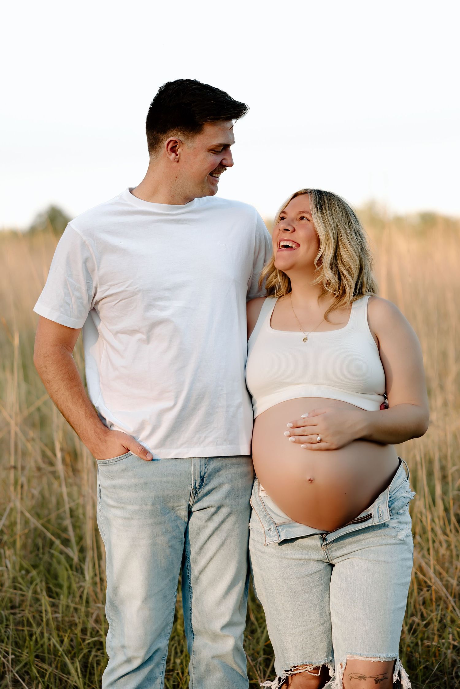 Couple in casual white and denim outfits pose together in a sunlit field during maternity photoshoot.