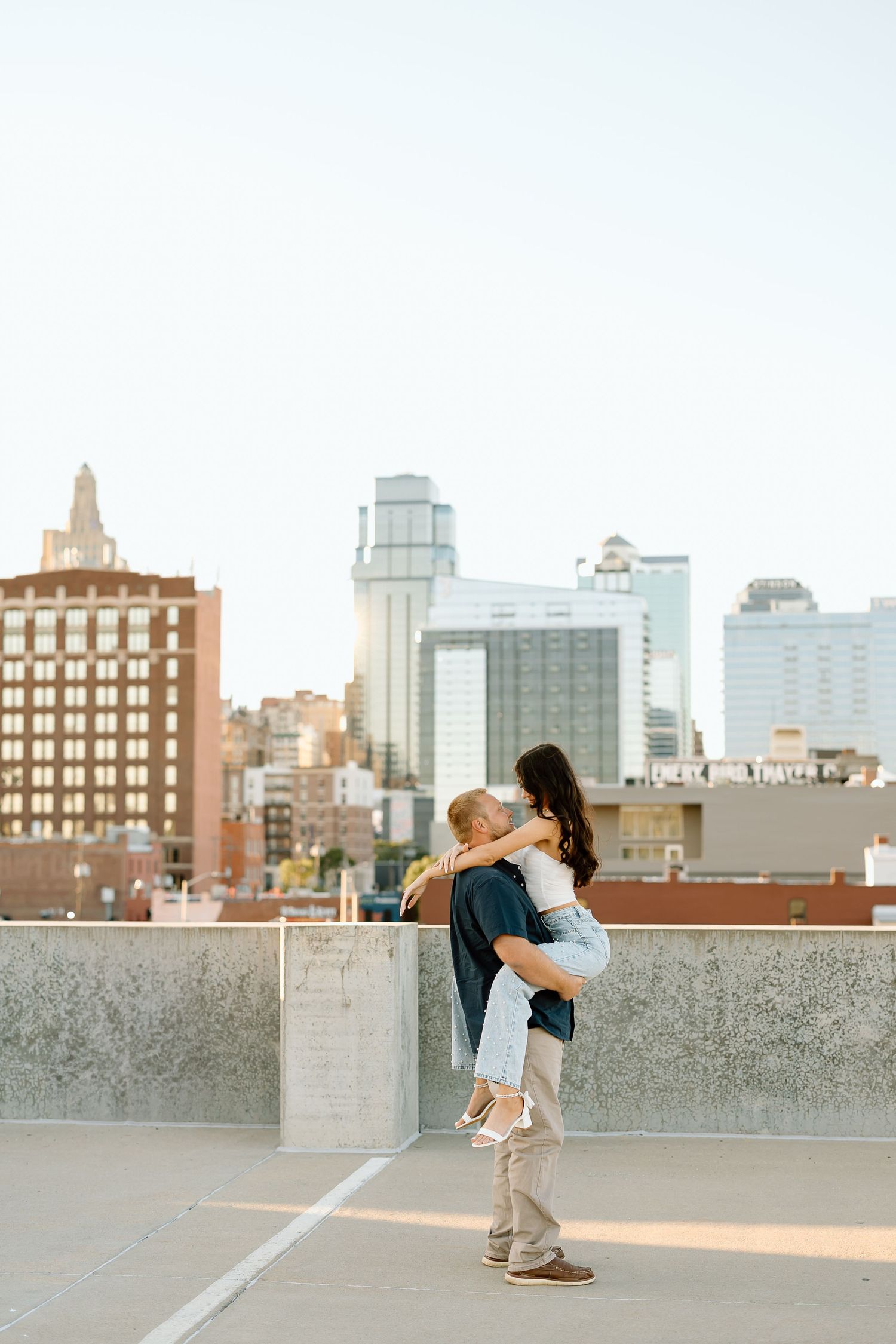 A romantic couple shares an intimate dance pose on a rooftop with an urban cityscape backdrop at sunset.