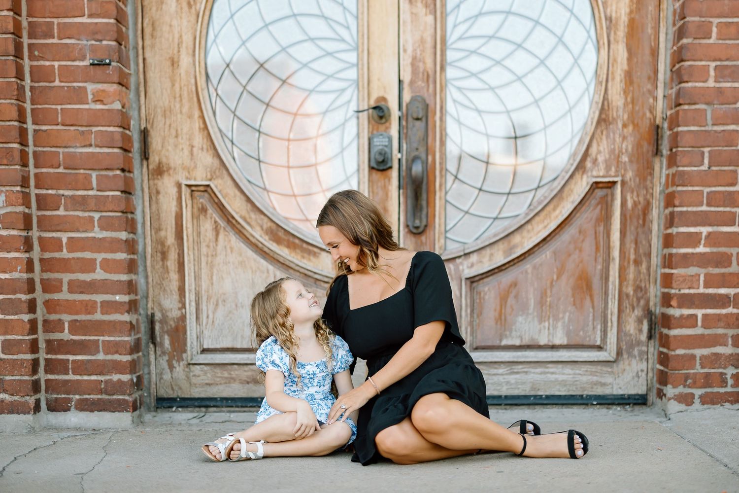 A person and child practice yoga stretches together on a wooden floor beside a brick wall with circular windows.