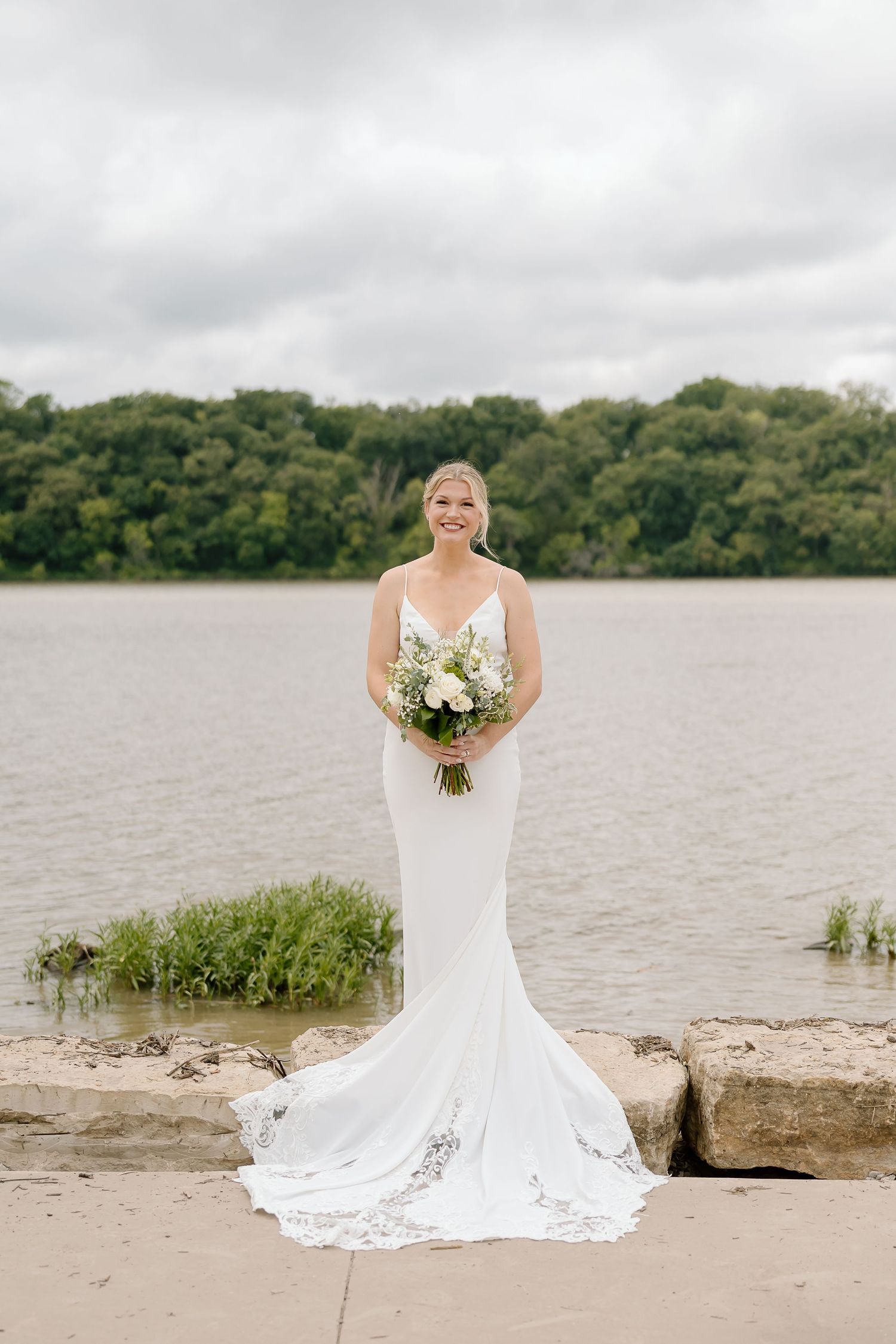A wedding portrait series at a lakeside beach with white dress and bouquet against a cloudy sky and wooded shore.