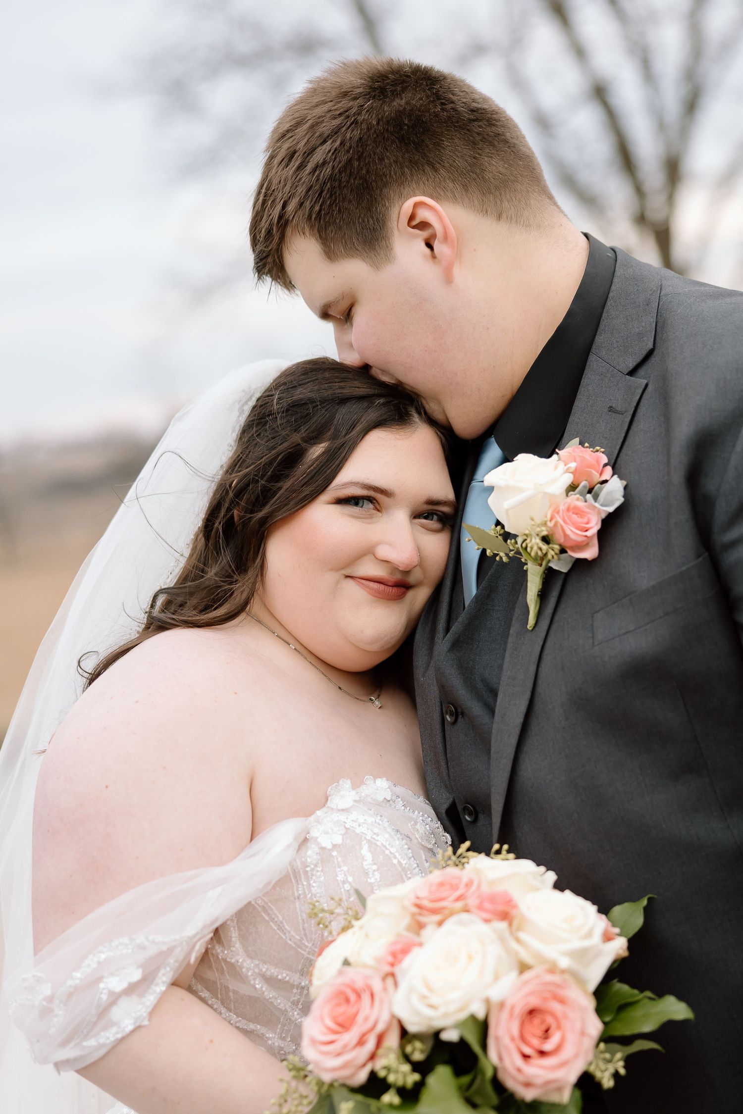 Romantic wedding couple poses together with coral and white rose bouquet and boutonniere in elegant attire.