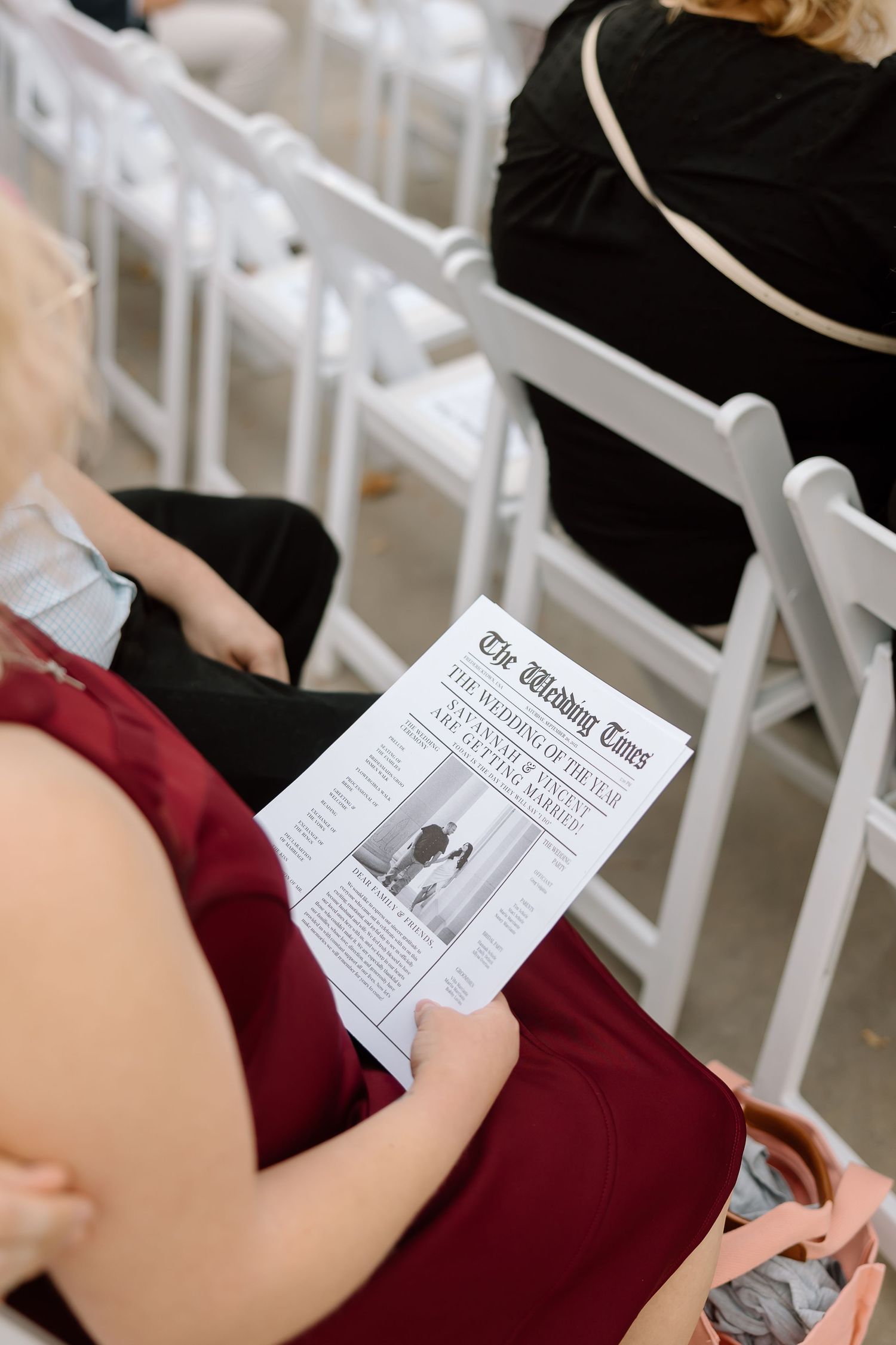 Wedding guest in burgundy dress reads program while seated in white folding chair at ceremony.