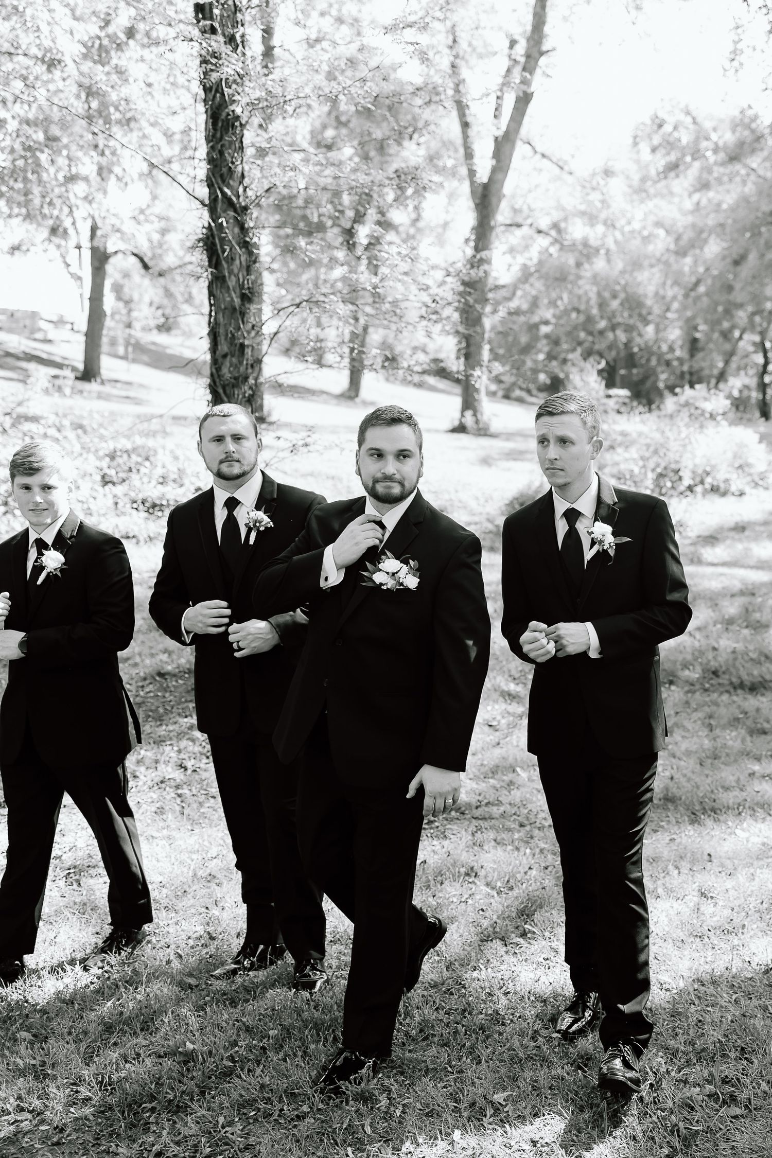 A black and white photo of groomsmen in suits posing playfully in a park setting.