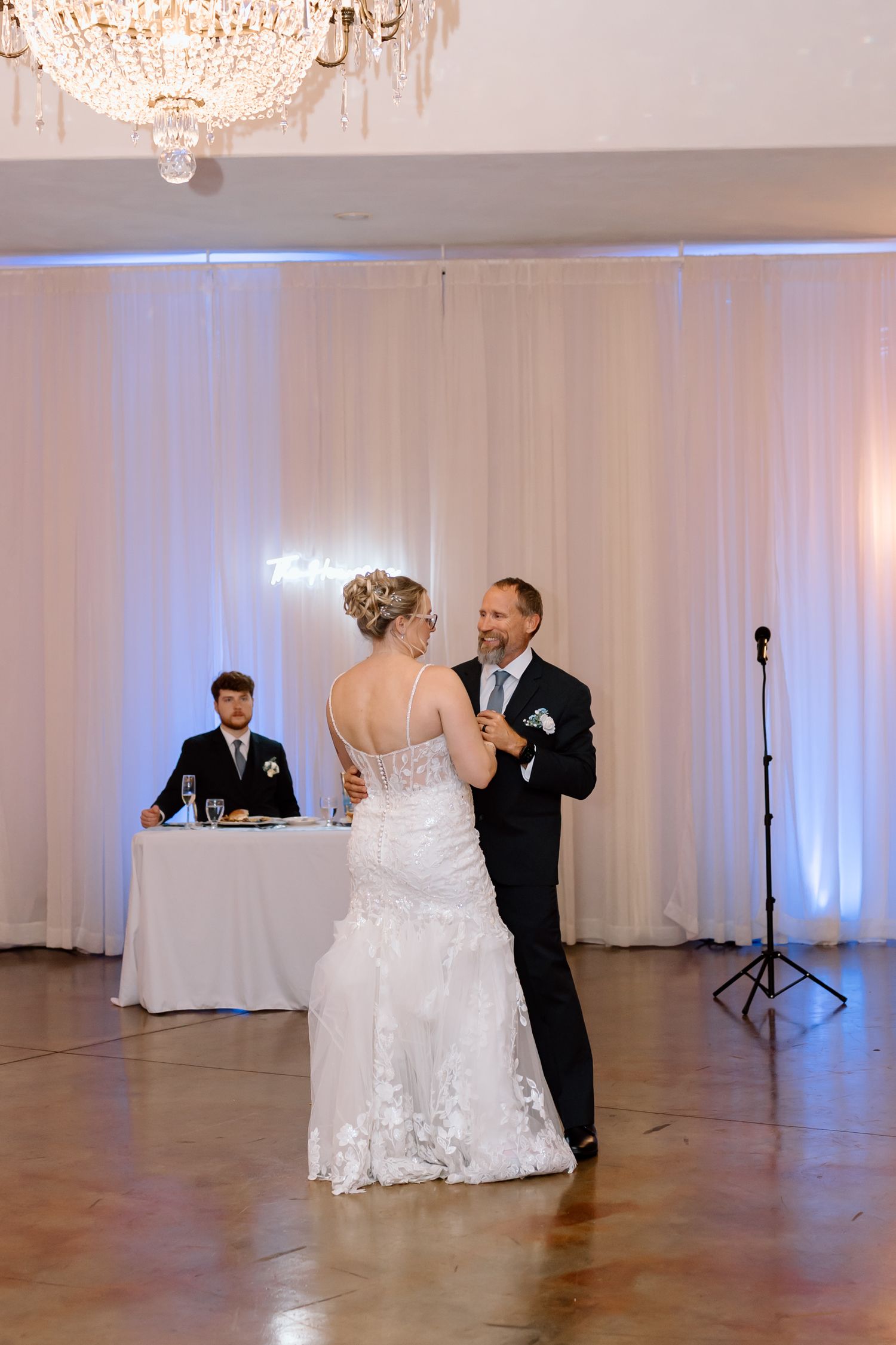 A couple shares their first dance at a wedding reception with a crystal chandelier overhead and white drapes in the background.