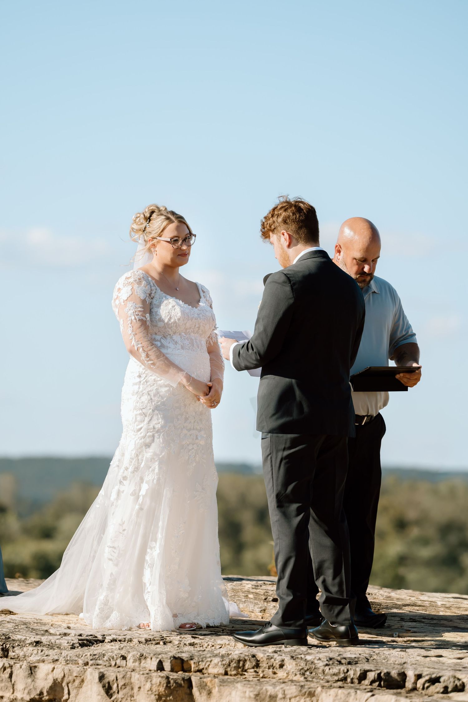 A couple in wedding attire stands together on a stone ledge overlooking a scenic landscape on their special day.