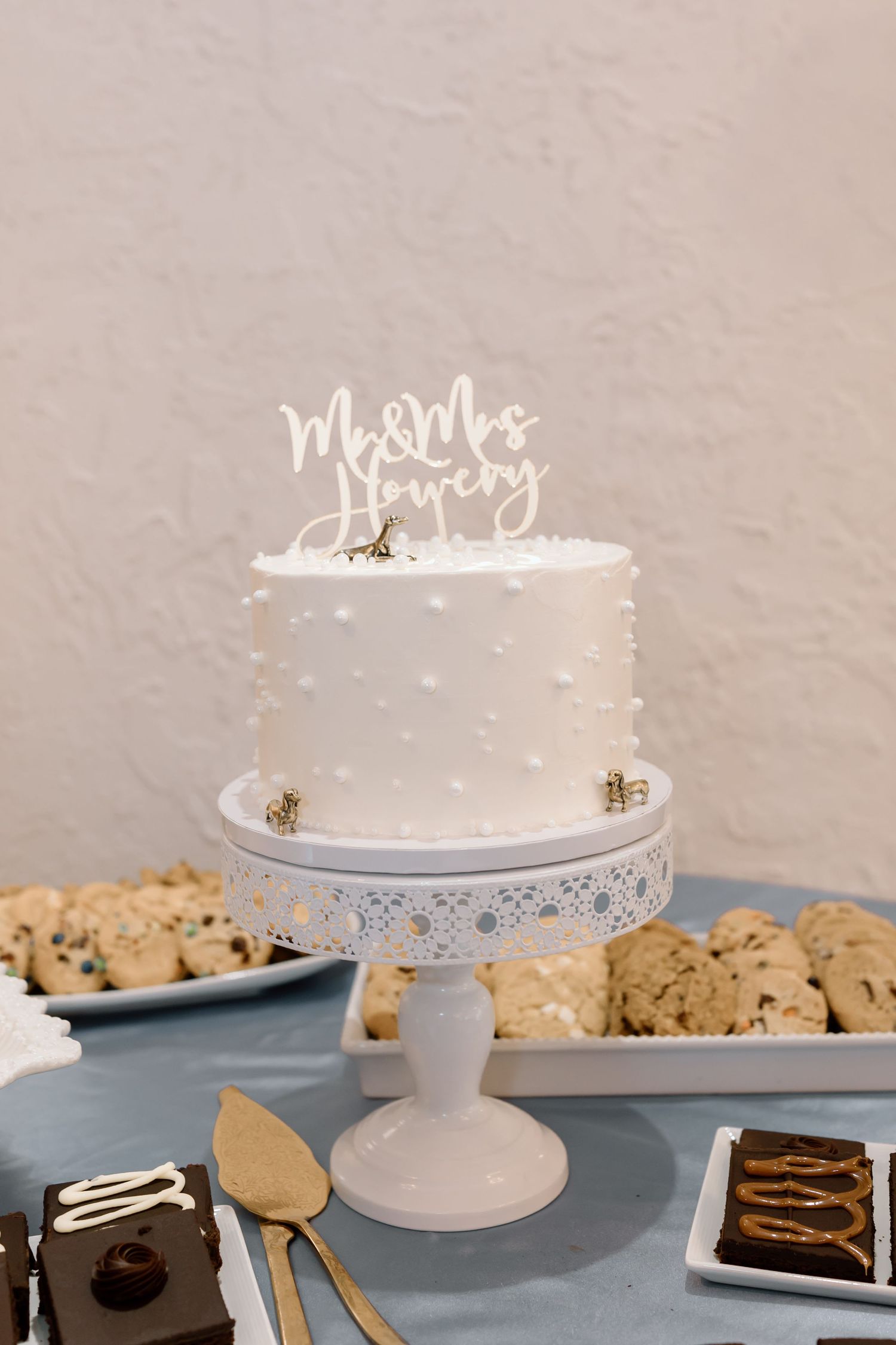 White wedding cake with polka dot texture and script topper displayed on ornate pedestal stand beside chocolate chip cookies.