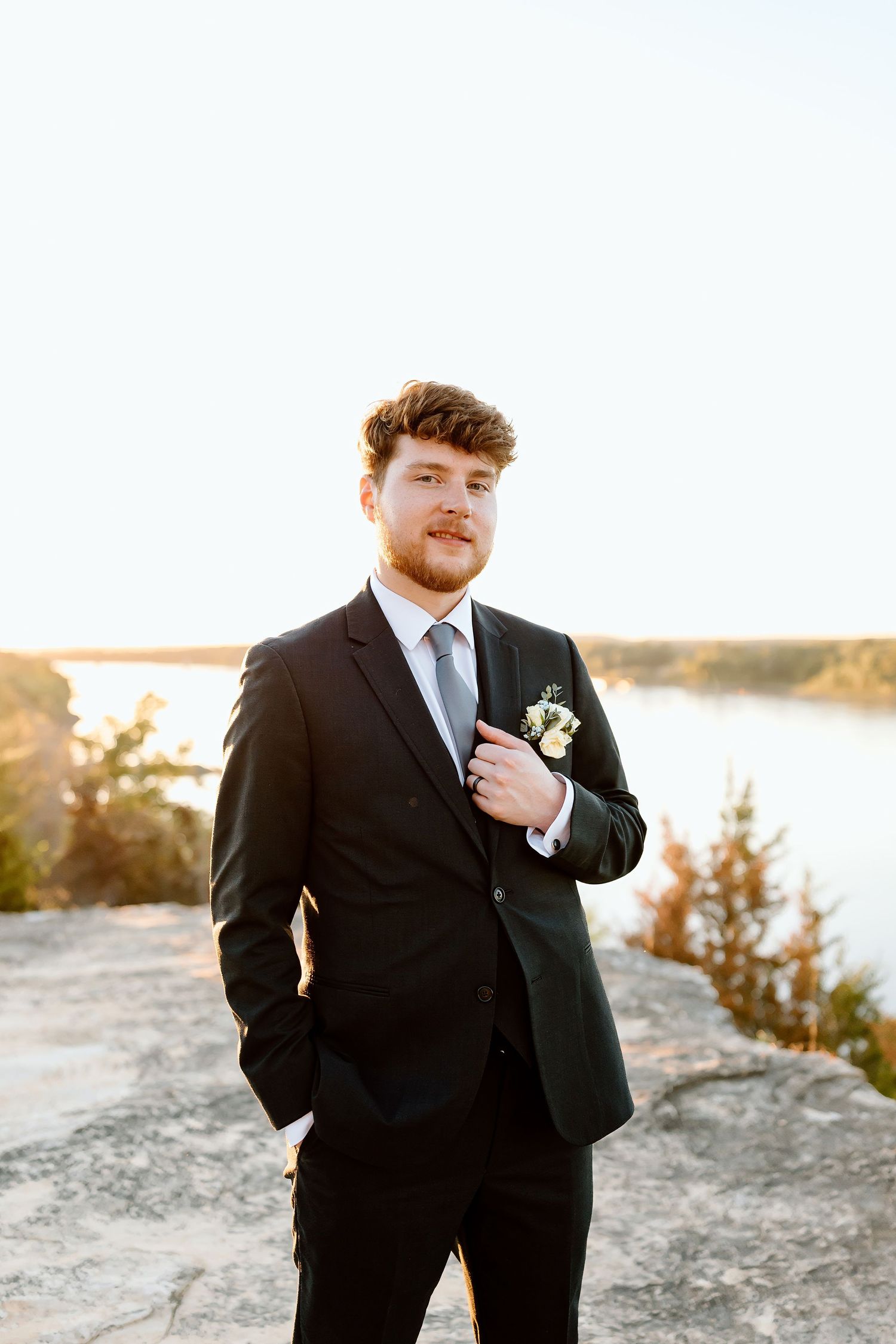 A groom in a black suit and blue tie poses on a rocky overlook at sunset with a boutonniere.