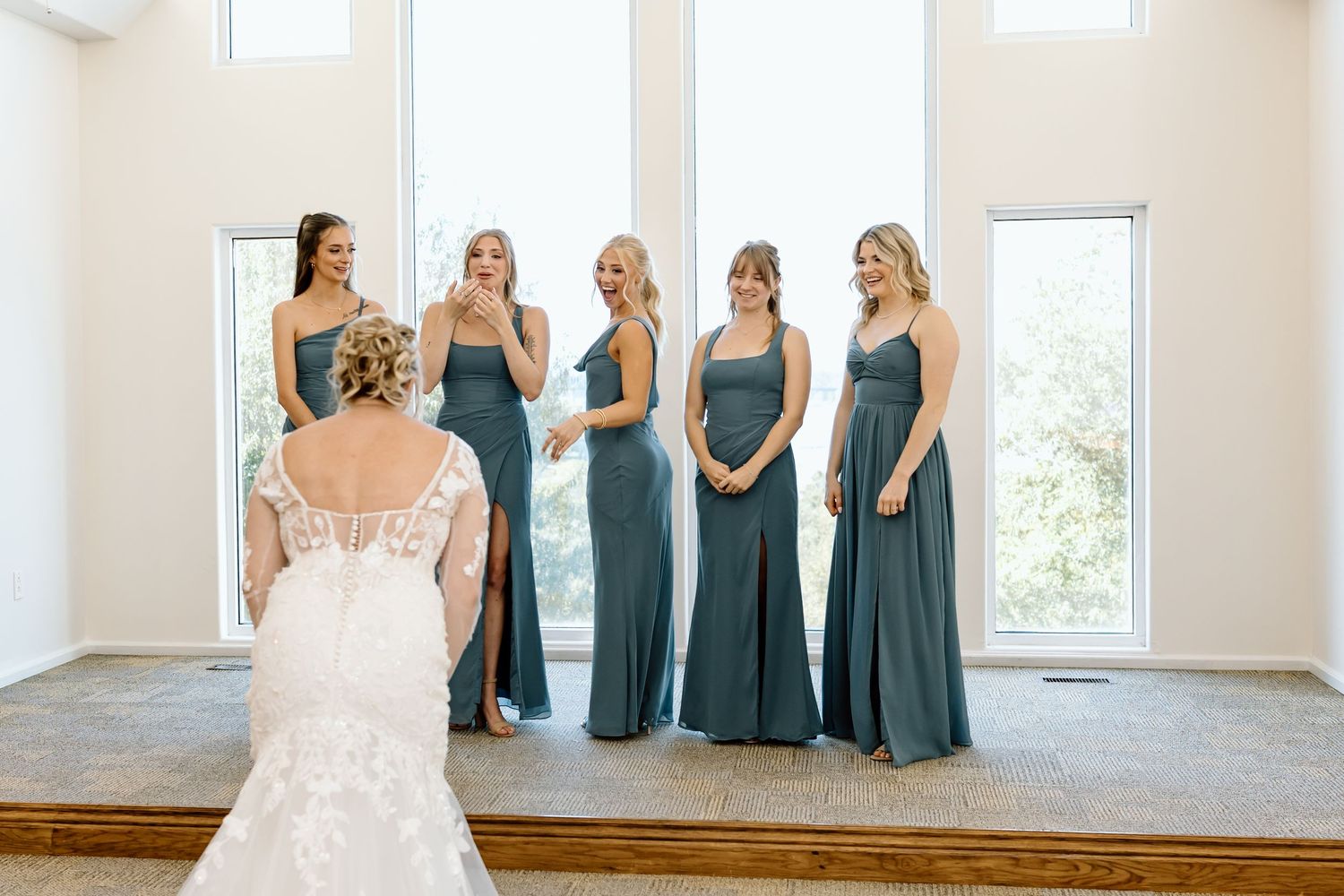 A group in matching dusty blue bridesmaid dresses stand together in a bright sunlit room during a wedding event.