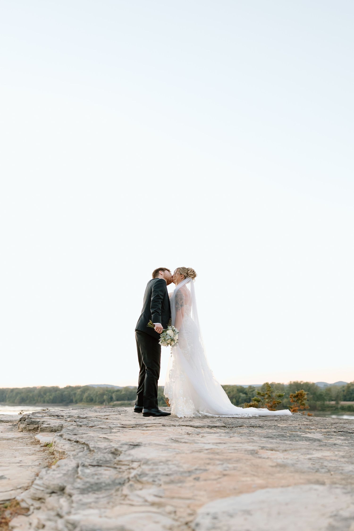 A couple shares an intimate moment on a rocky cliff overlooking a scenic landscape during their wedding photoshoot.