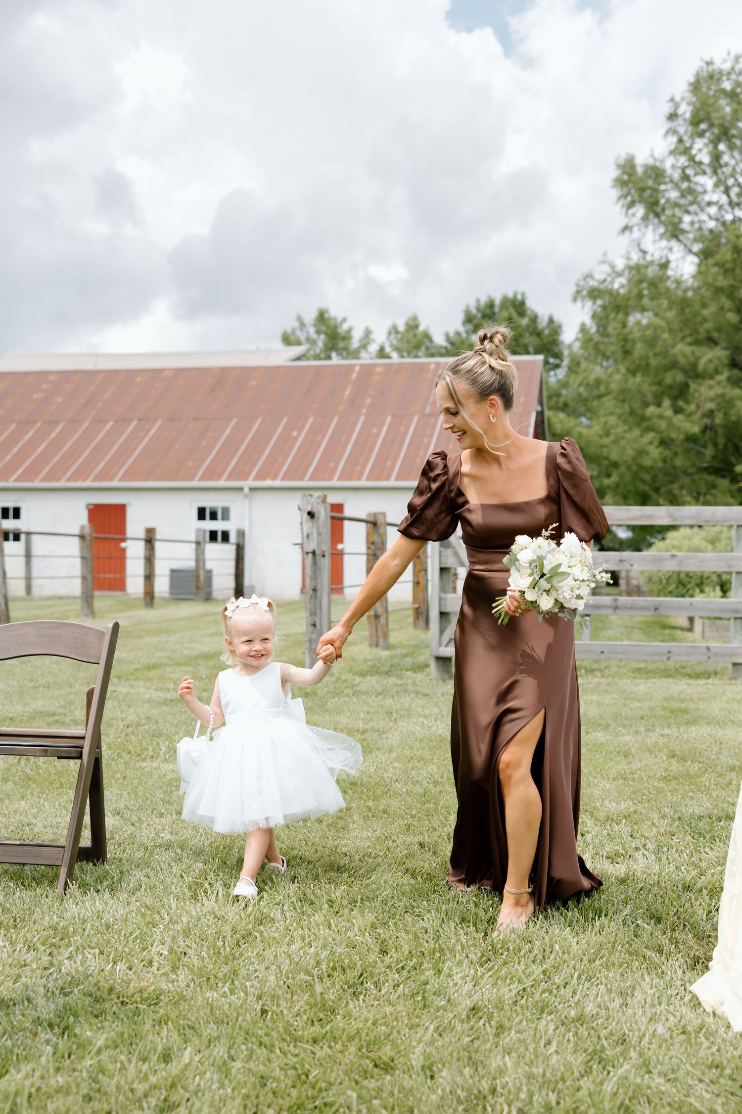 A bridesmaid in a brown satin dress holds hands with a flower girl in white tulle at an outdoor farm wedding.