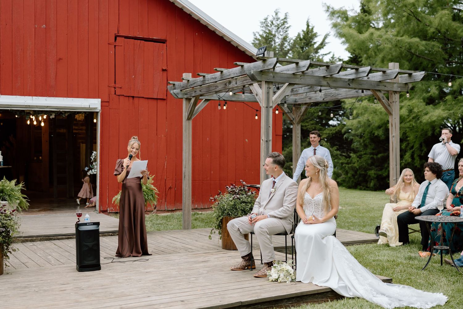 Outdoor barn wedding ceremony on a wooden deck with bride and groom seated before a pergola beside a red barn.