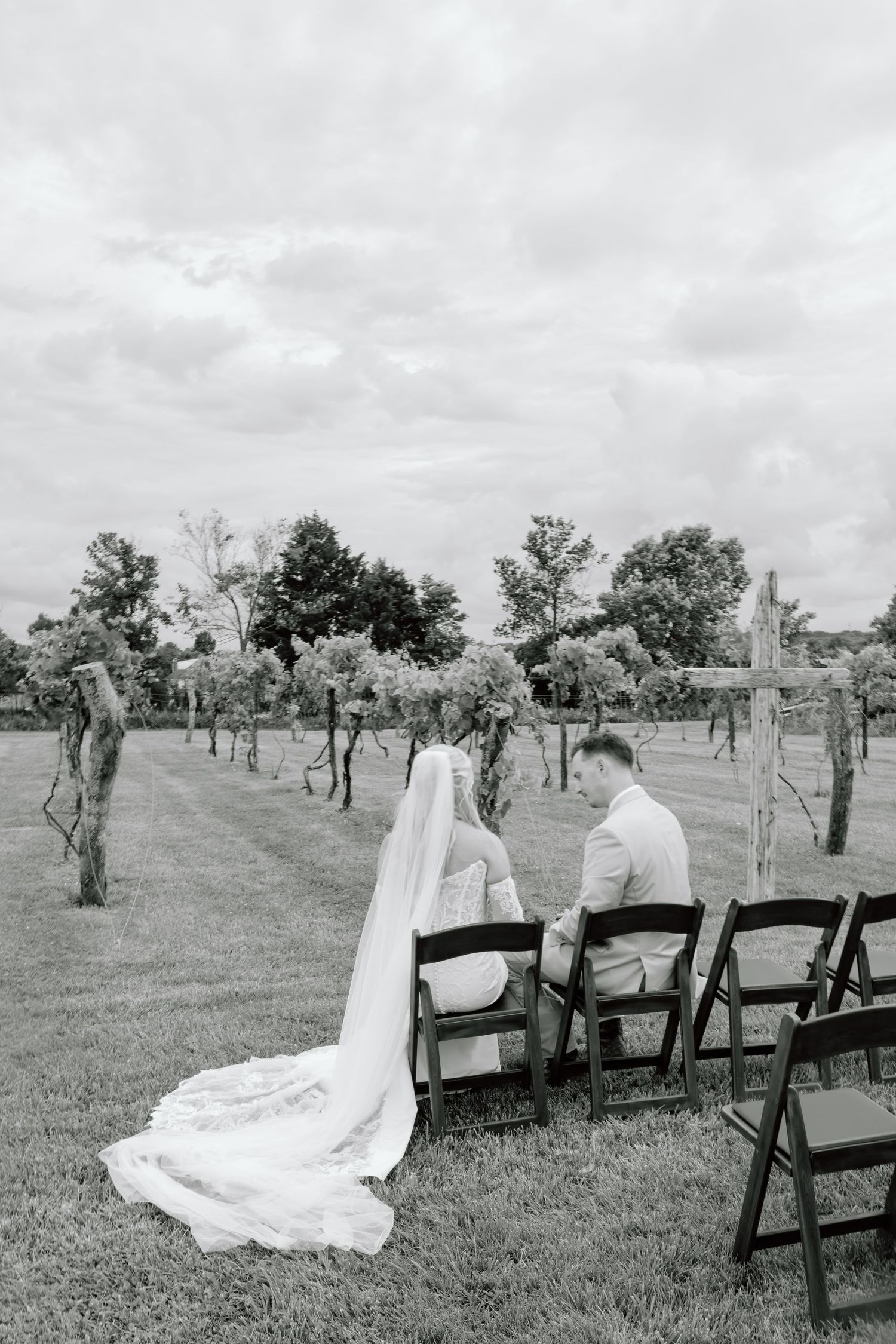 Black and white photo of bride and groom sitting in chairs at outdoor vineyard wedding ceremony.