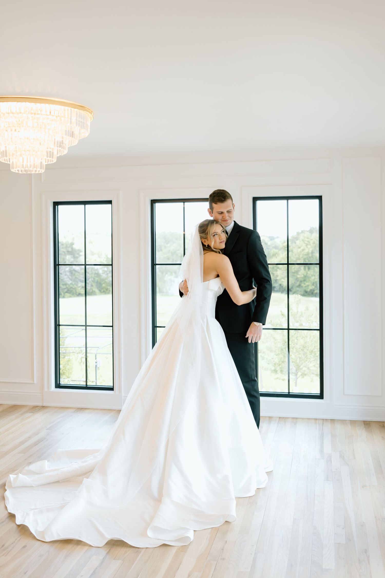 Bride and groom share a romantic embrace in an elegant white room with black-framed windows and a crystal chandelier.