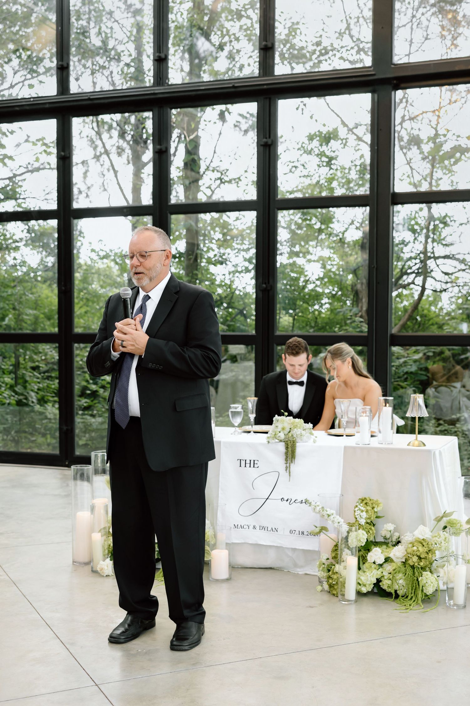Older man in black suit giving wedding toast at reception, bride and groom seated at sweetheart table behind him.