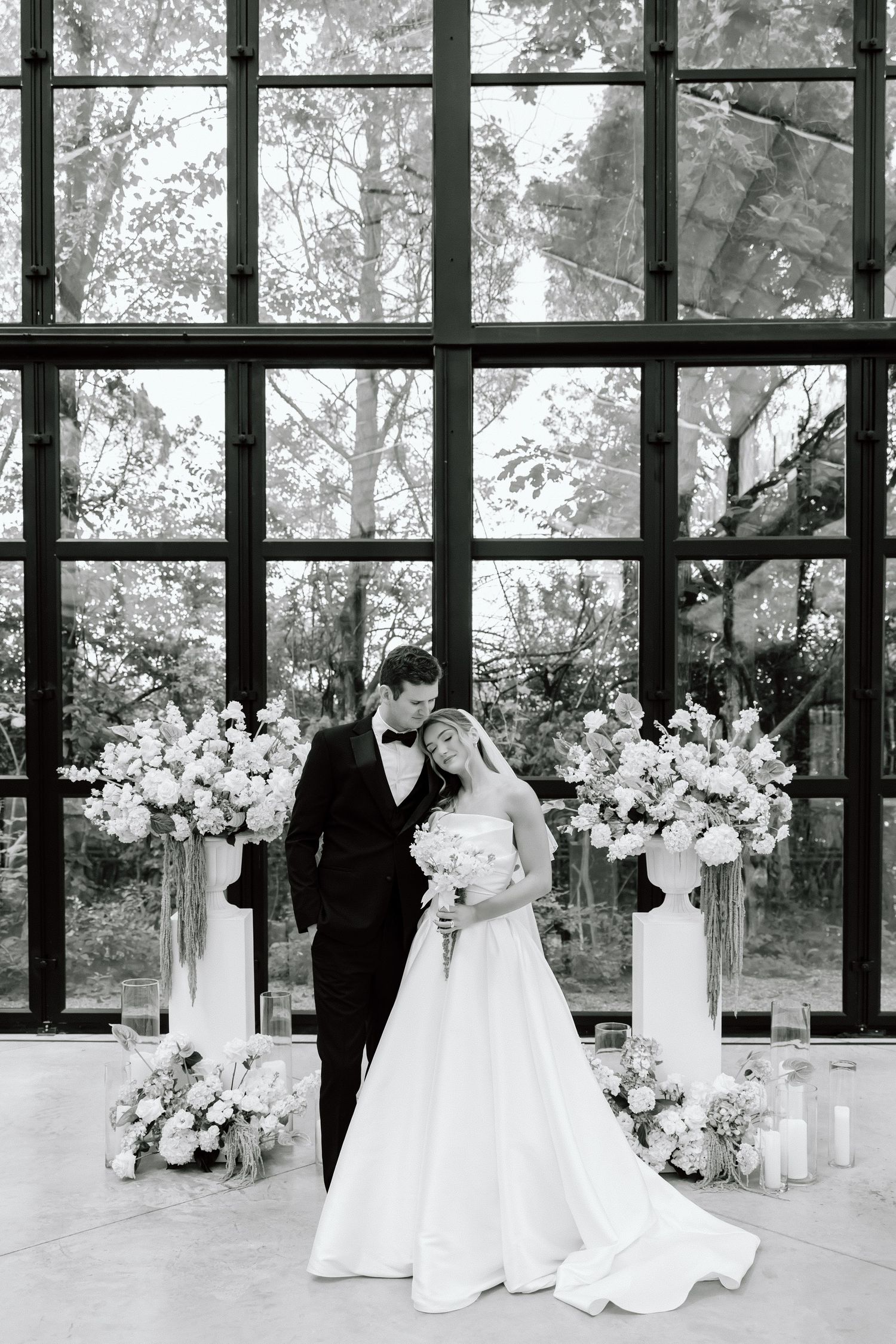 Black and white wedding portrait of bride and groom embracing before large windows with elegant floral arrangements.