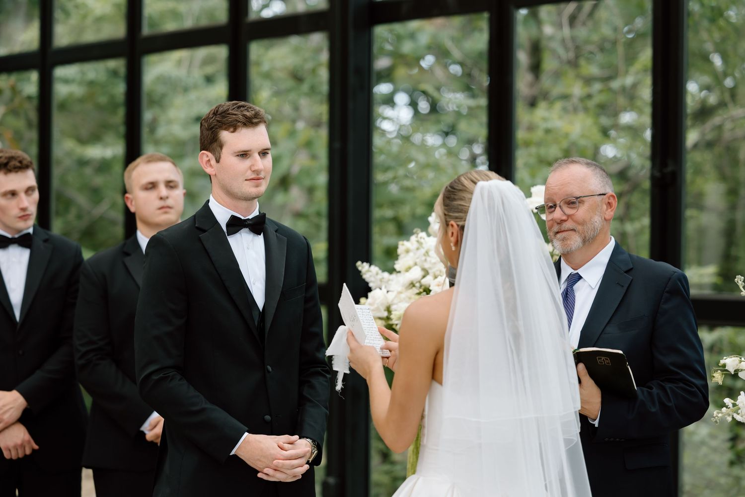 Groom in black tuxedo stands at wedding altar with groomsmen while bride holds white bouquet during ceremony.