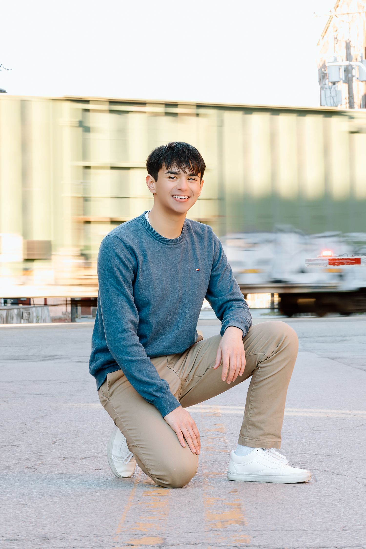 Candid portrait of person in blue sweater kneeling on concrete in industrial urban setting.