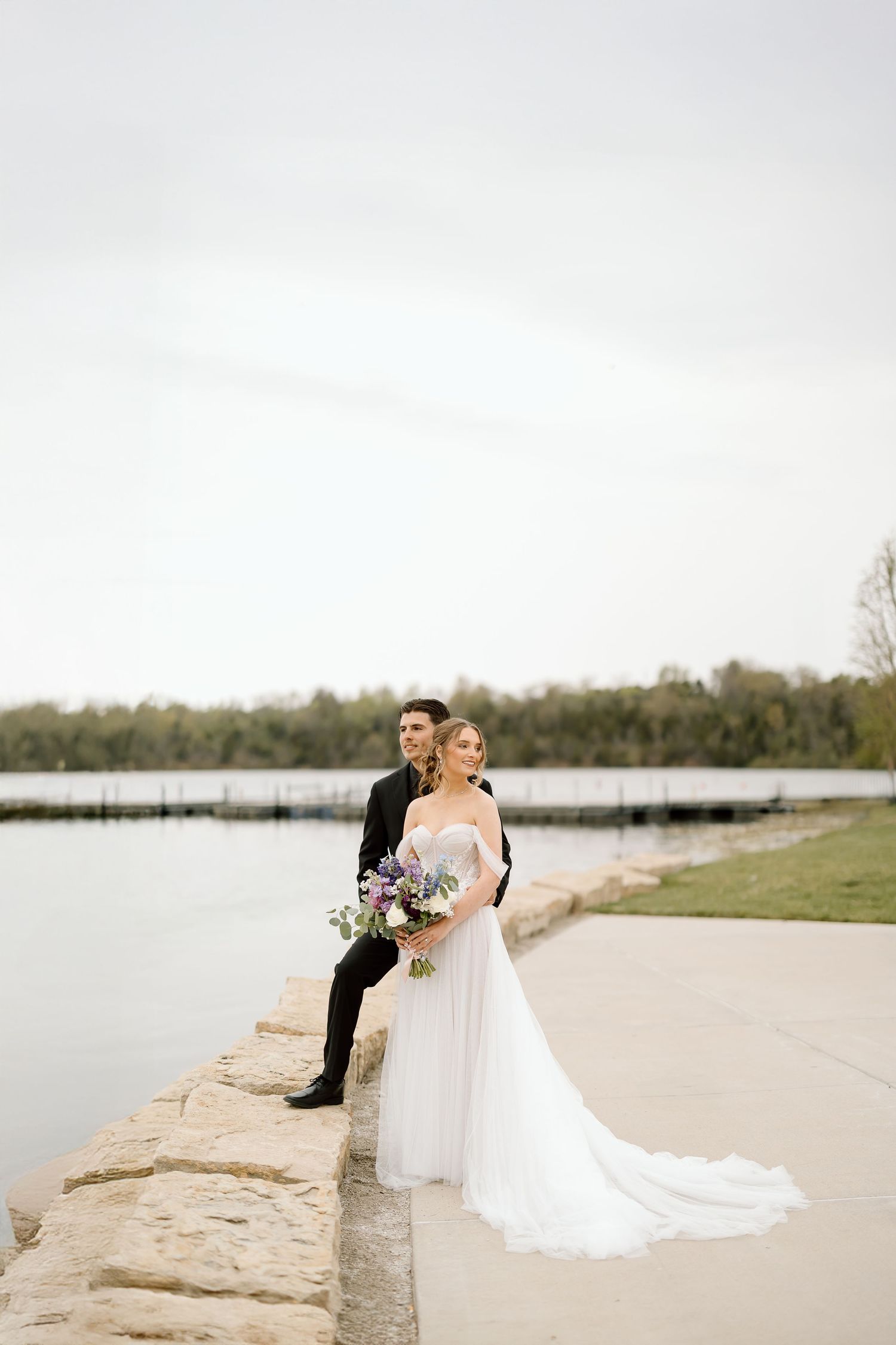 Wedding couple poses on lakeside stone ledge with bride holding floral bouquet.