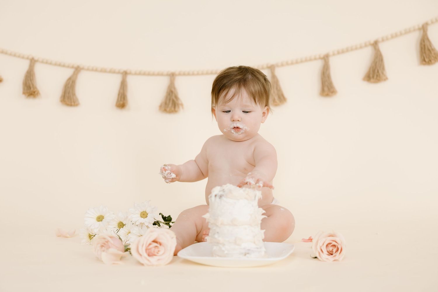Small child interacting with white cake surrounded by roses against beige background with tassels.