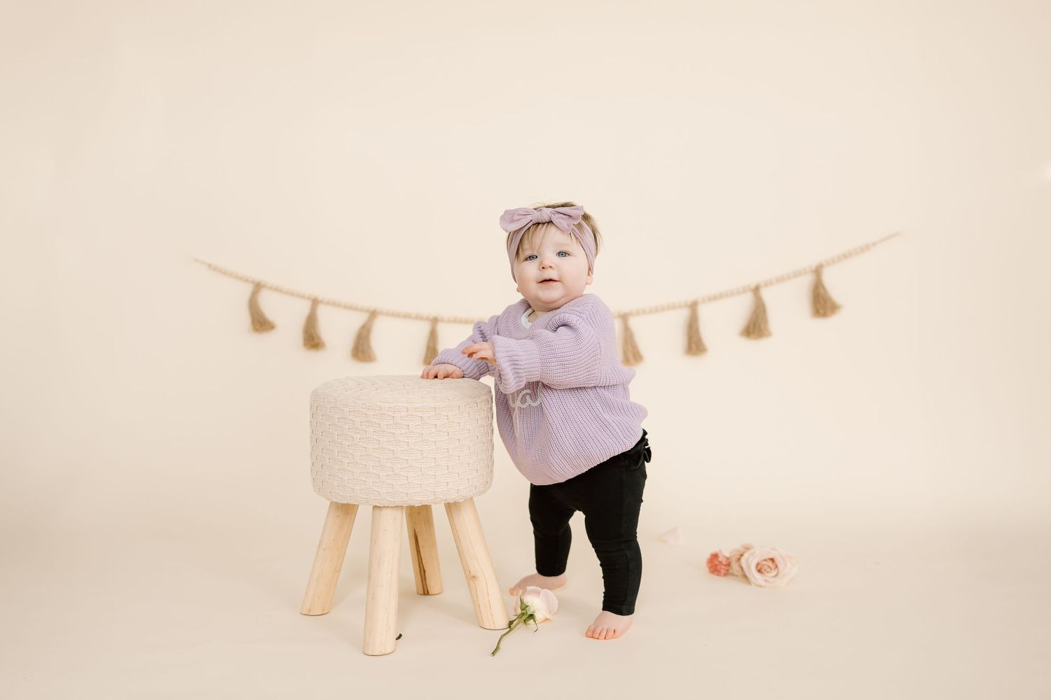 Small child in purple sweater stands near wooden stool under hanging tassel garland.