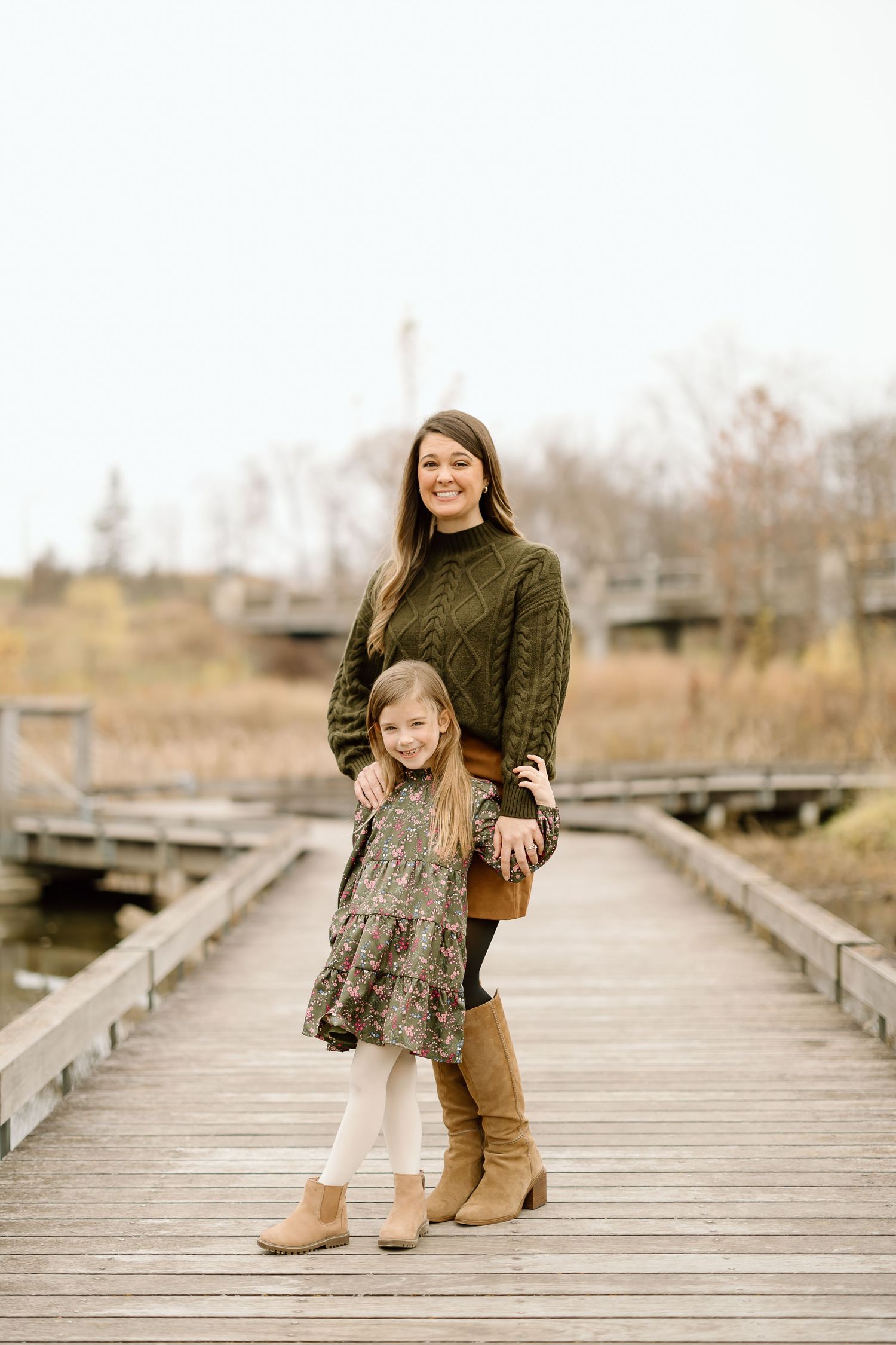 A parent and child stand together on a wooden boardwalk wearing coordinated autumn outfits.