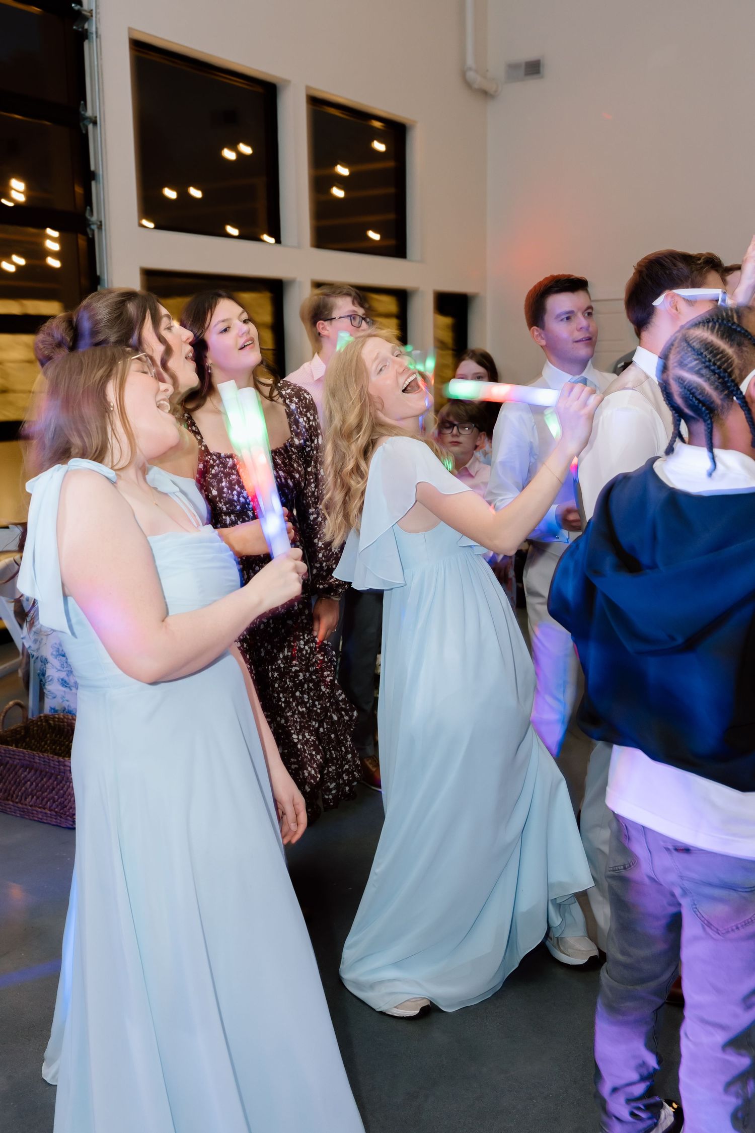 Bridesmaids in light blue gowns dancing and laughing with glow sticks at a lively wedding reception.