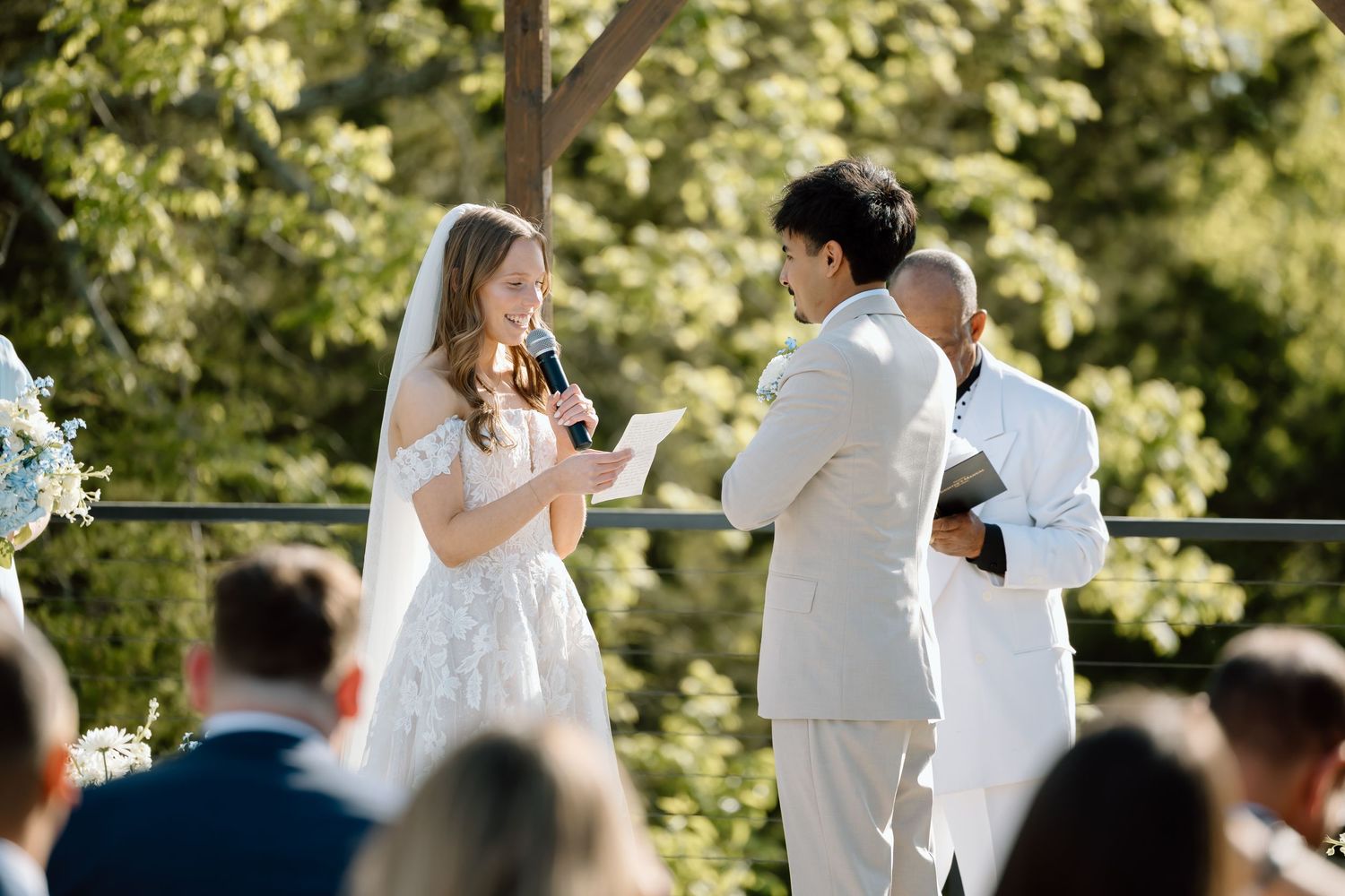 Bride reads vows to groom at outdoor wedding ceremony while guests watch and officiant stands nearby.
