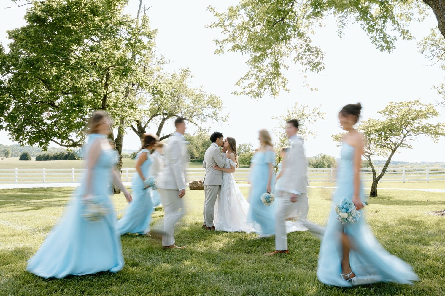 Wedding party in light blue dresses walks across green lawn as bride and groom kiss.