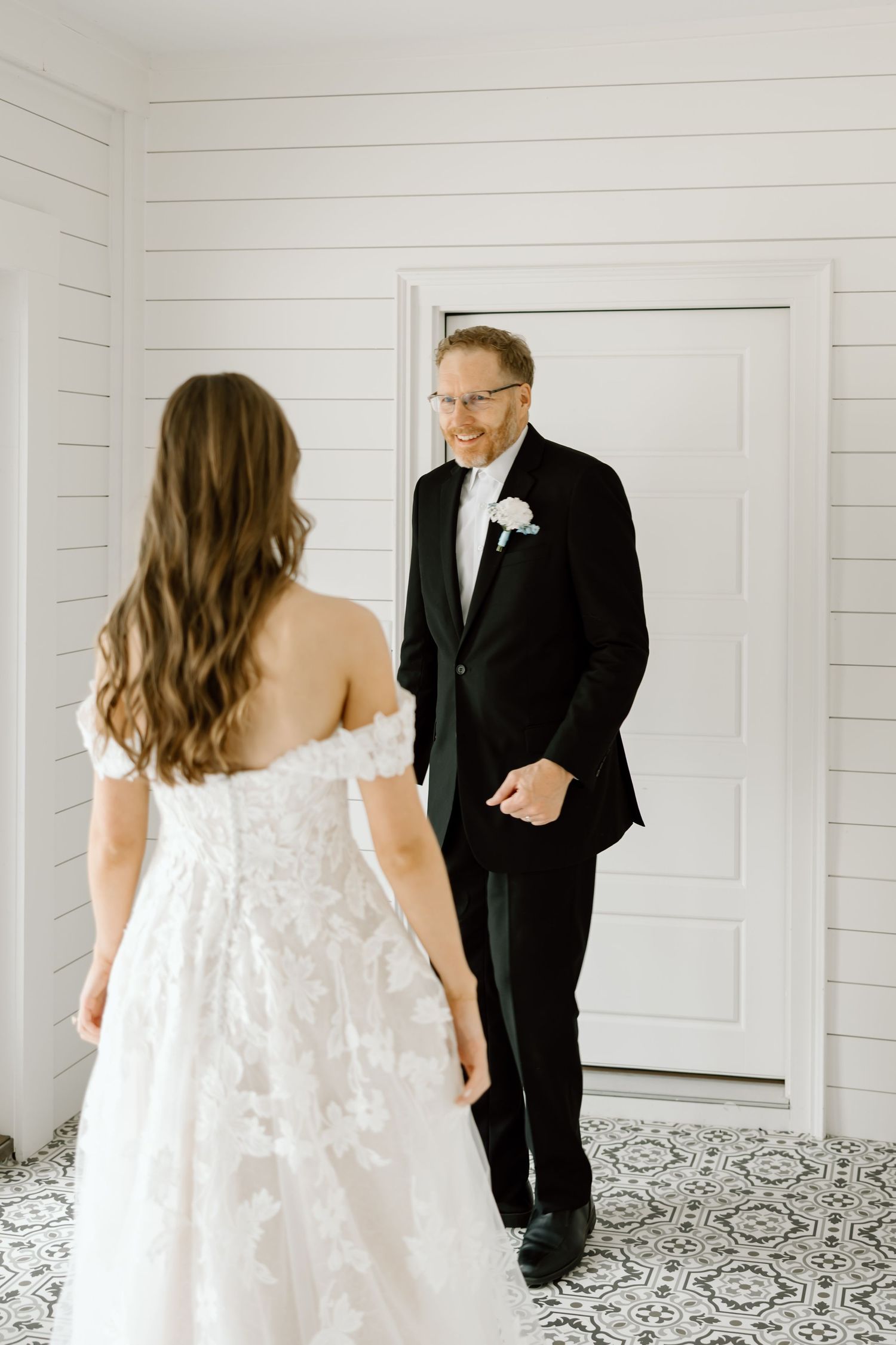 A bride in a lace gown shares a first look moment with a smiling groom in a black tuxedo.