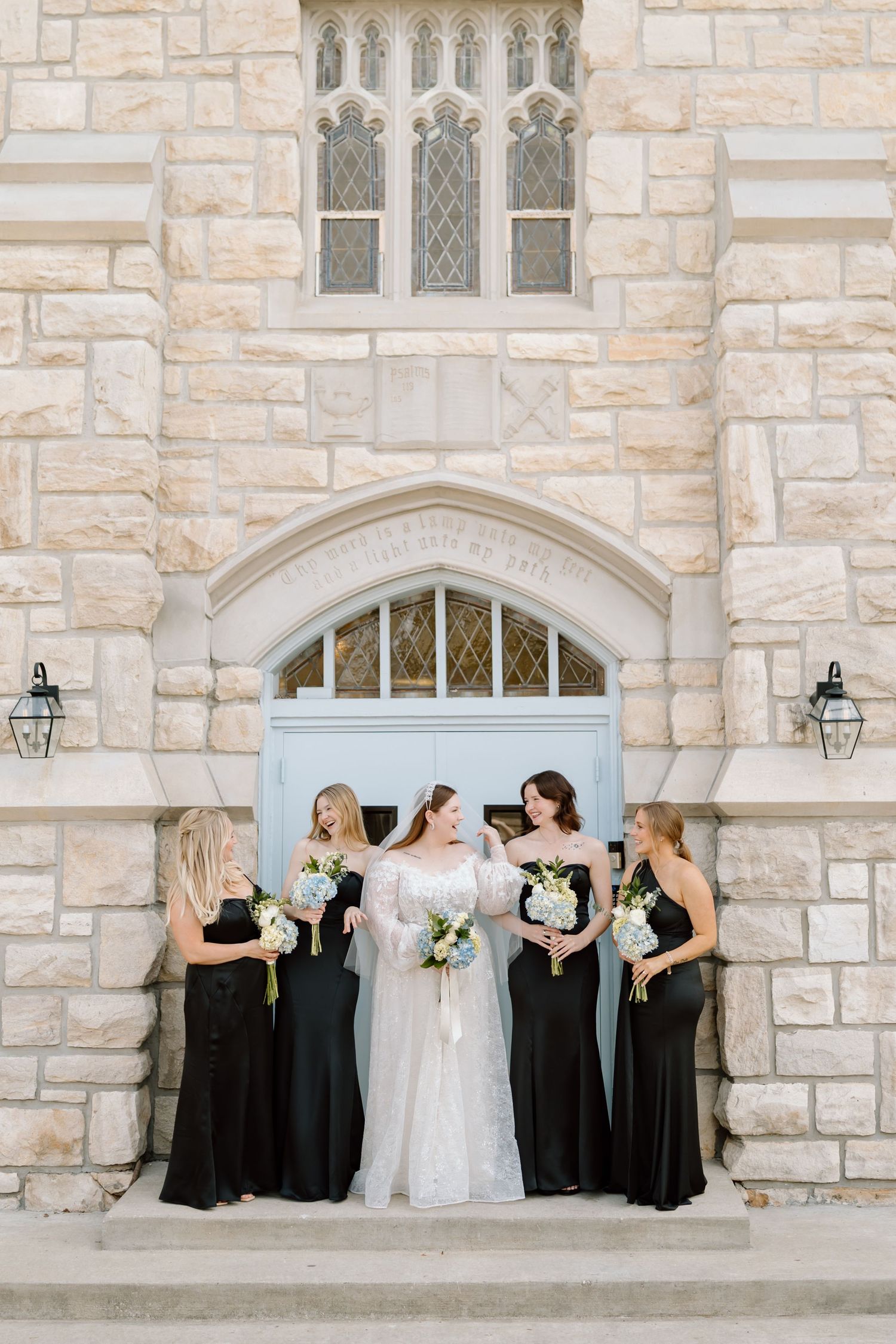 Bride in lace gown stands with four bridesmaids in black dresses holding bouquets before a stone church blue door.