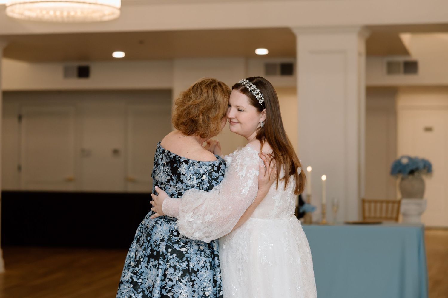 A bride in a white lace gown embraces a woman in a floral dress at a wedding reception.