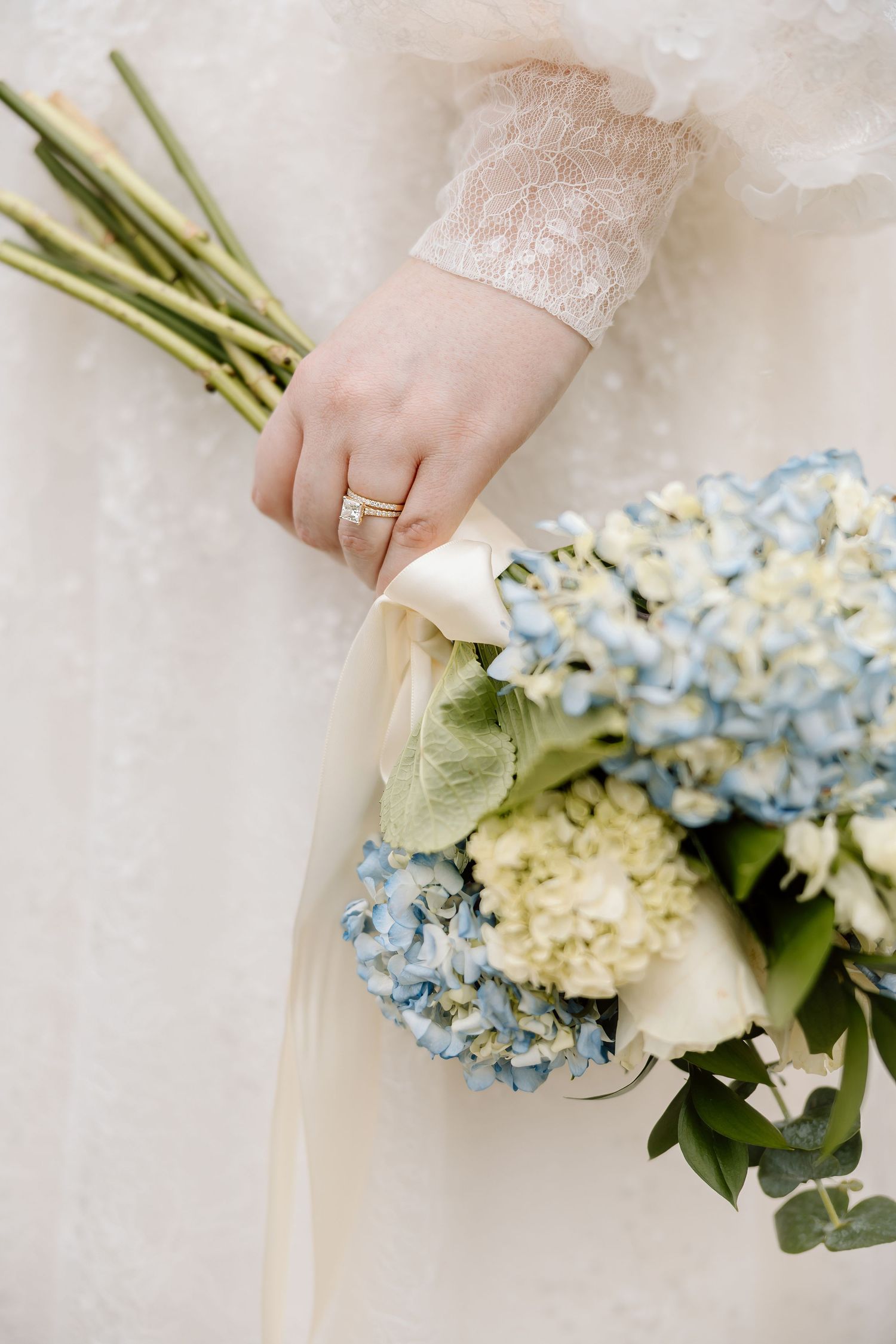 Bride holding blue and white hydrangea wedding bouquet with ribbon, wearing lace dress and gold ring.