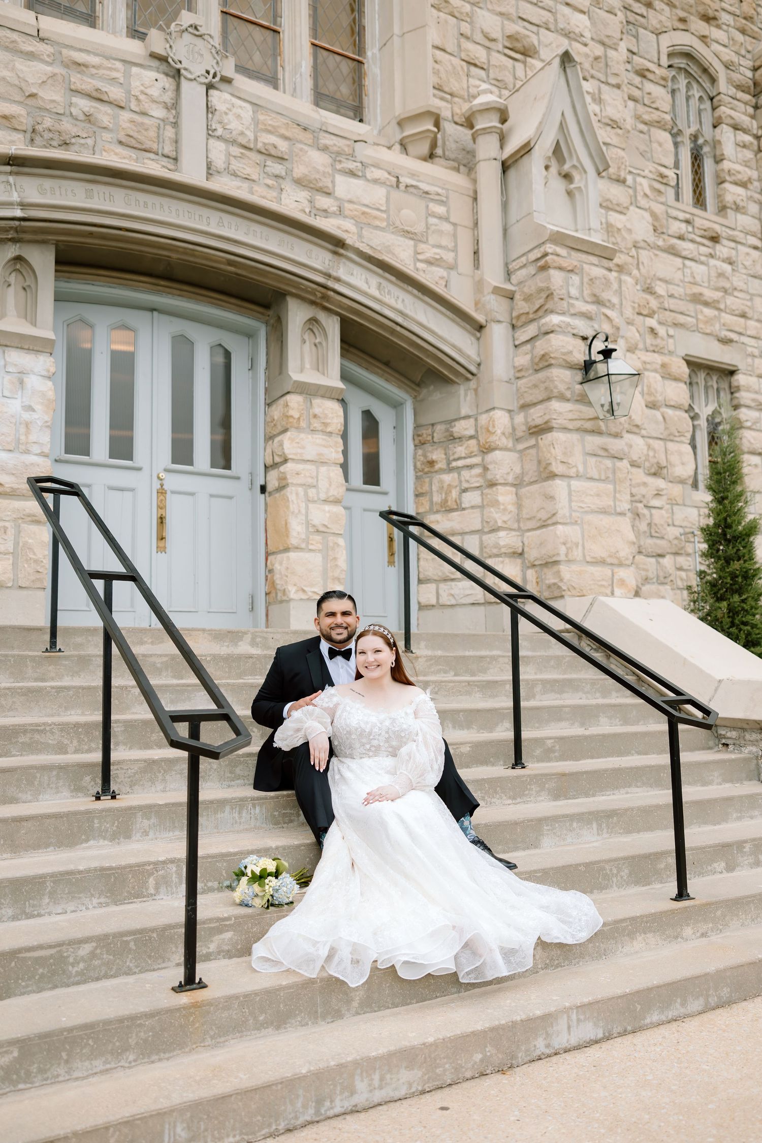 Bride and groom pose on stone church steps, she in a flowing lace gown, he in a black tuxedo, holding a white bouquet.