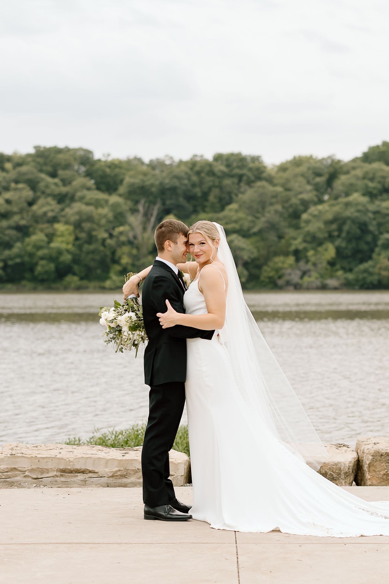 A romantic wedding couple shares an intimate moment on a lakeside stone path in matching formal attire.