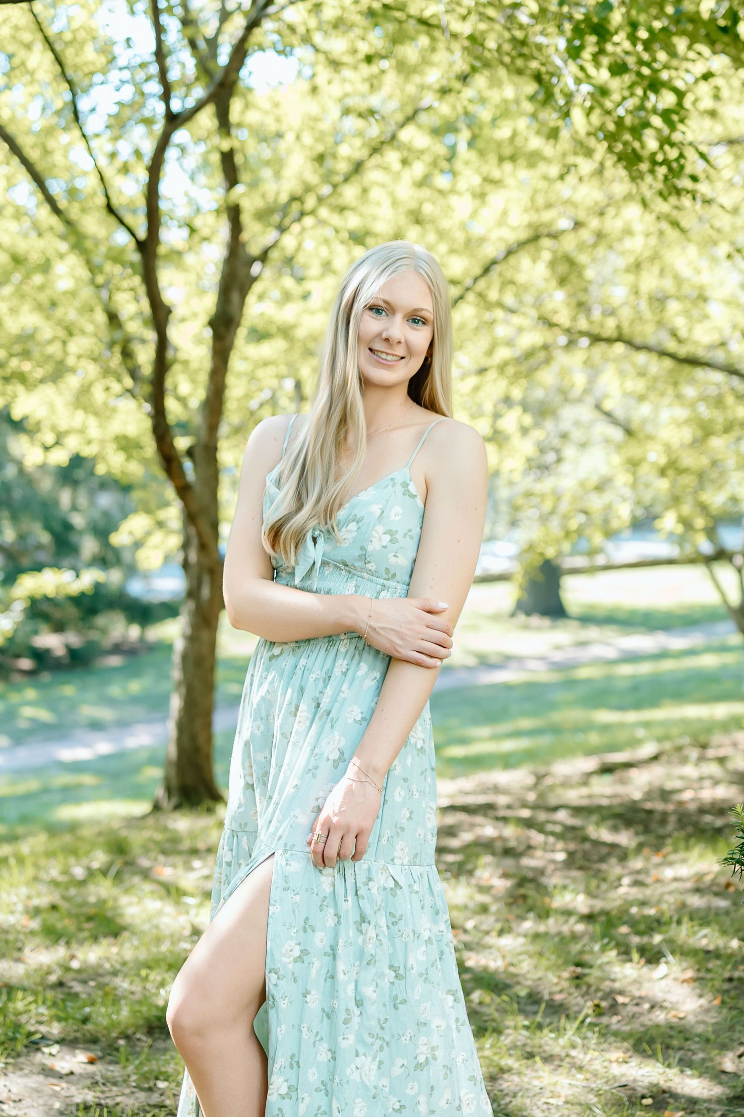 A person in a light blue floral sundress poses outdoors in a sunlit wooded area during a summer photo session.