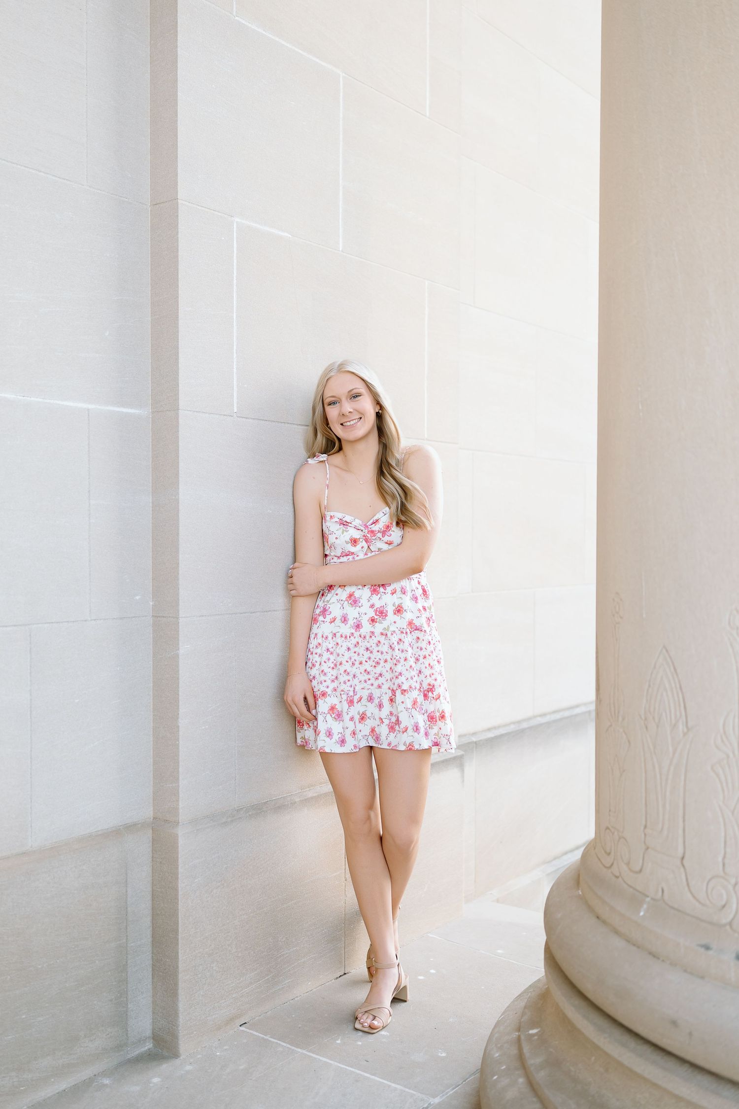 A person in a floral dress poses gracefully against white columns in a bright, minimalist architectural setting.