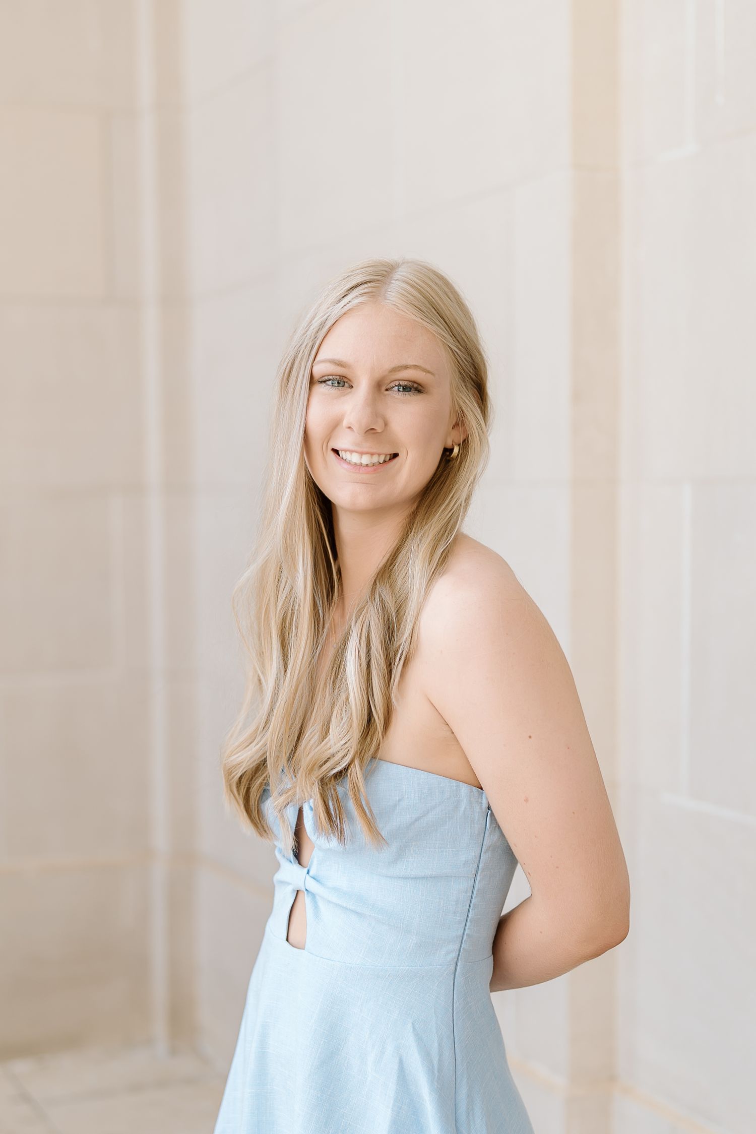 A person wearing a light blue sleeveless dress poses and smiles in a bright interior setting.
