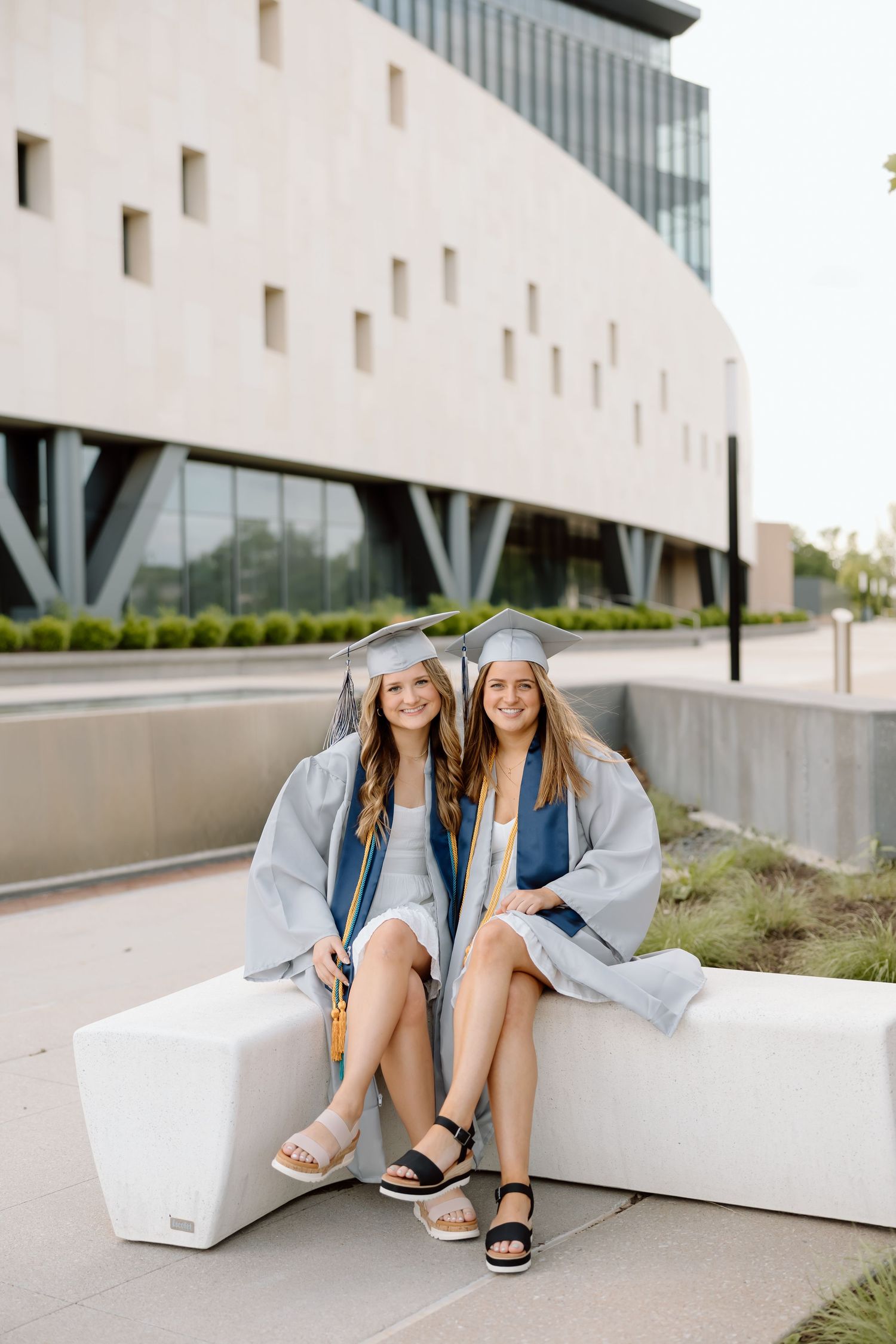 Two graduates in matching caps and gowns sit together on white concrete steps during a graduation photoshoot.