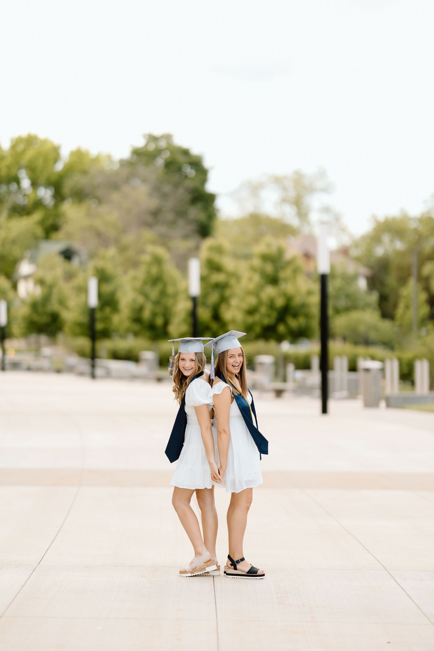 Two graduates in white dresses and navy cardigans stand back to back on campus plaza during graduation photoshoot.